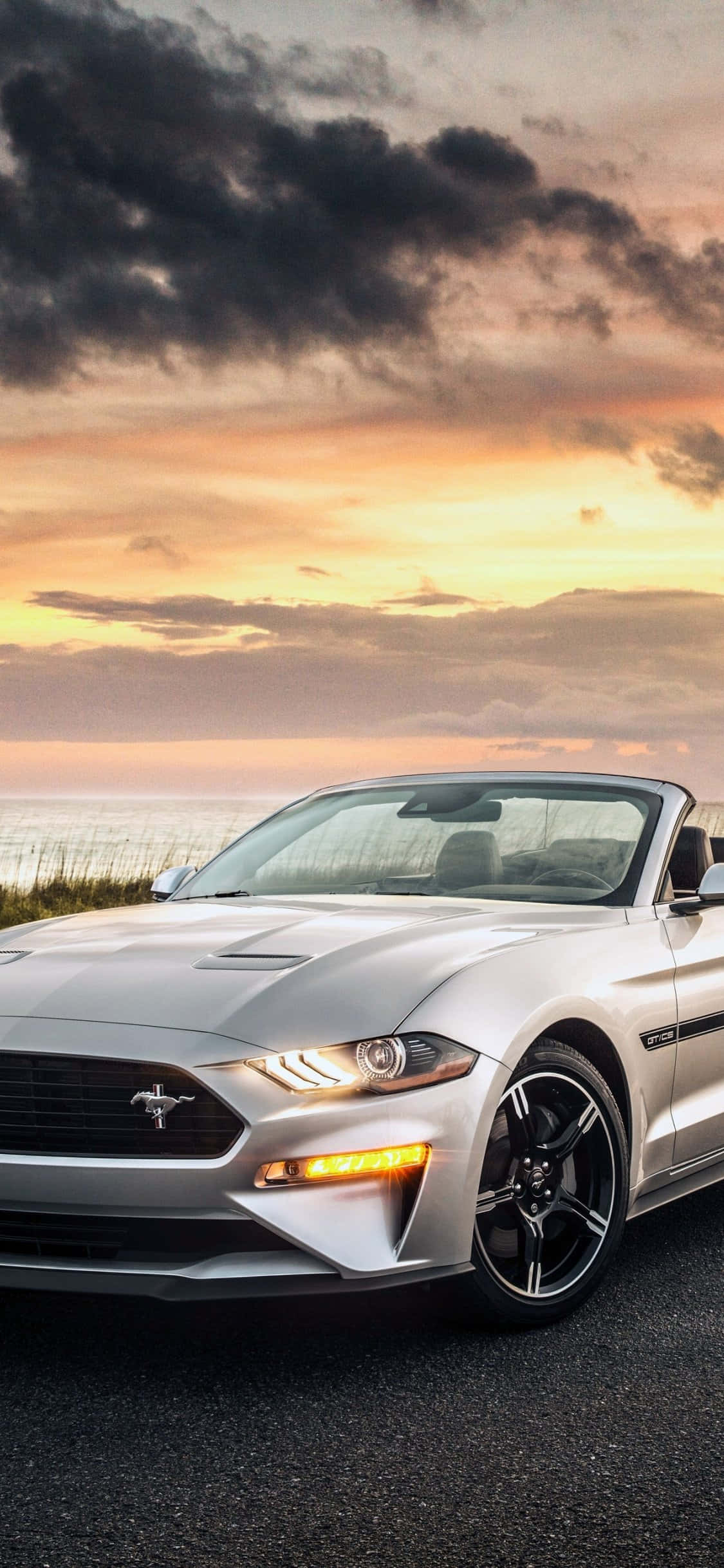 Mustang iPhone 桌布 壁紙 — 一輛銀色敞篷跑車停泊在海岸公路上，天空呈現戲劇性的日落景色，帶有橙色和紫色的雲彩。