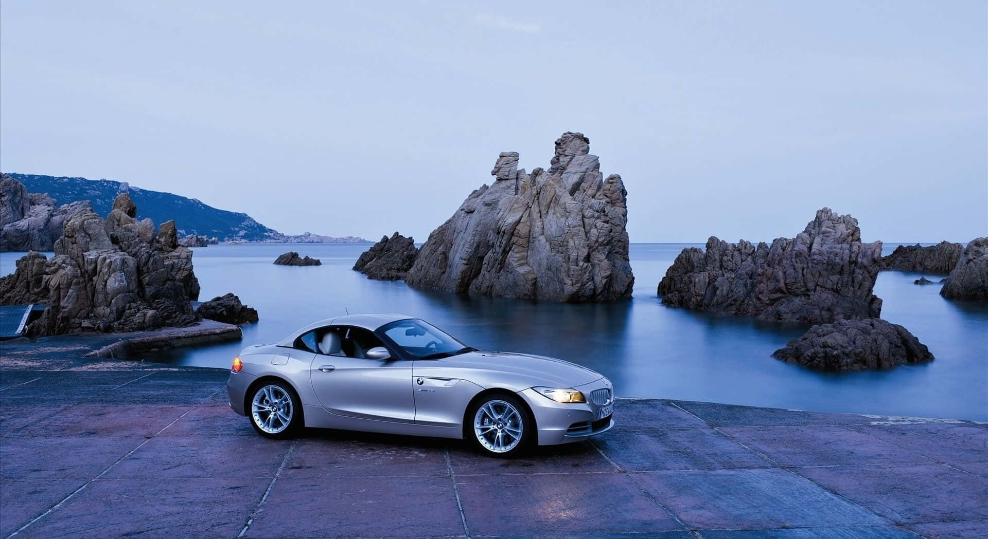 Bmw Z4 桌布 壁紙 — 一輛銀色敞篷跑車停泊在石碼頭旁，旁邊是平靜的海水，黃昏時分有戲劇性的岩石 formations。
