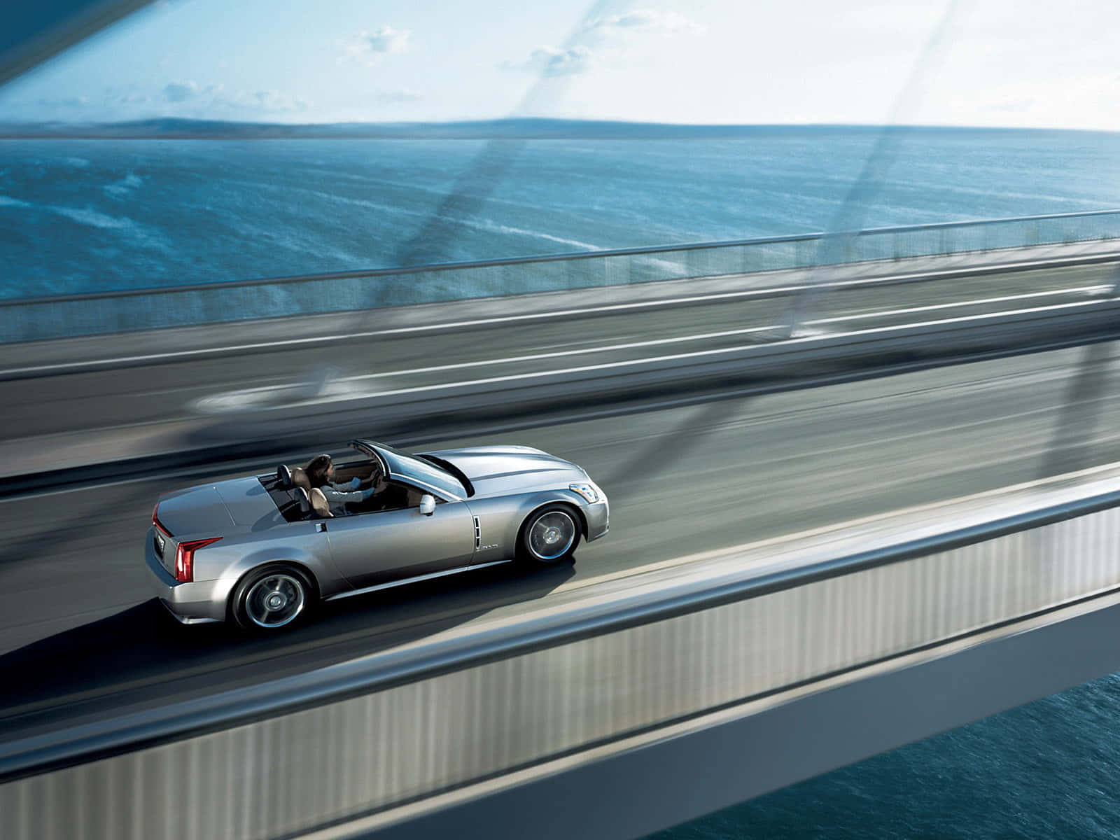 Cadillac Xlr 桌布 壁紙 — 一輛銀色敞篷跑車在晴朗天空下，沿著一座可俯瞰廣闊藍色海洋的現代化橋樑飛馳。