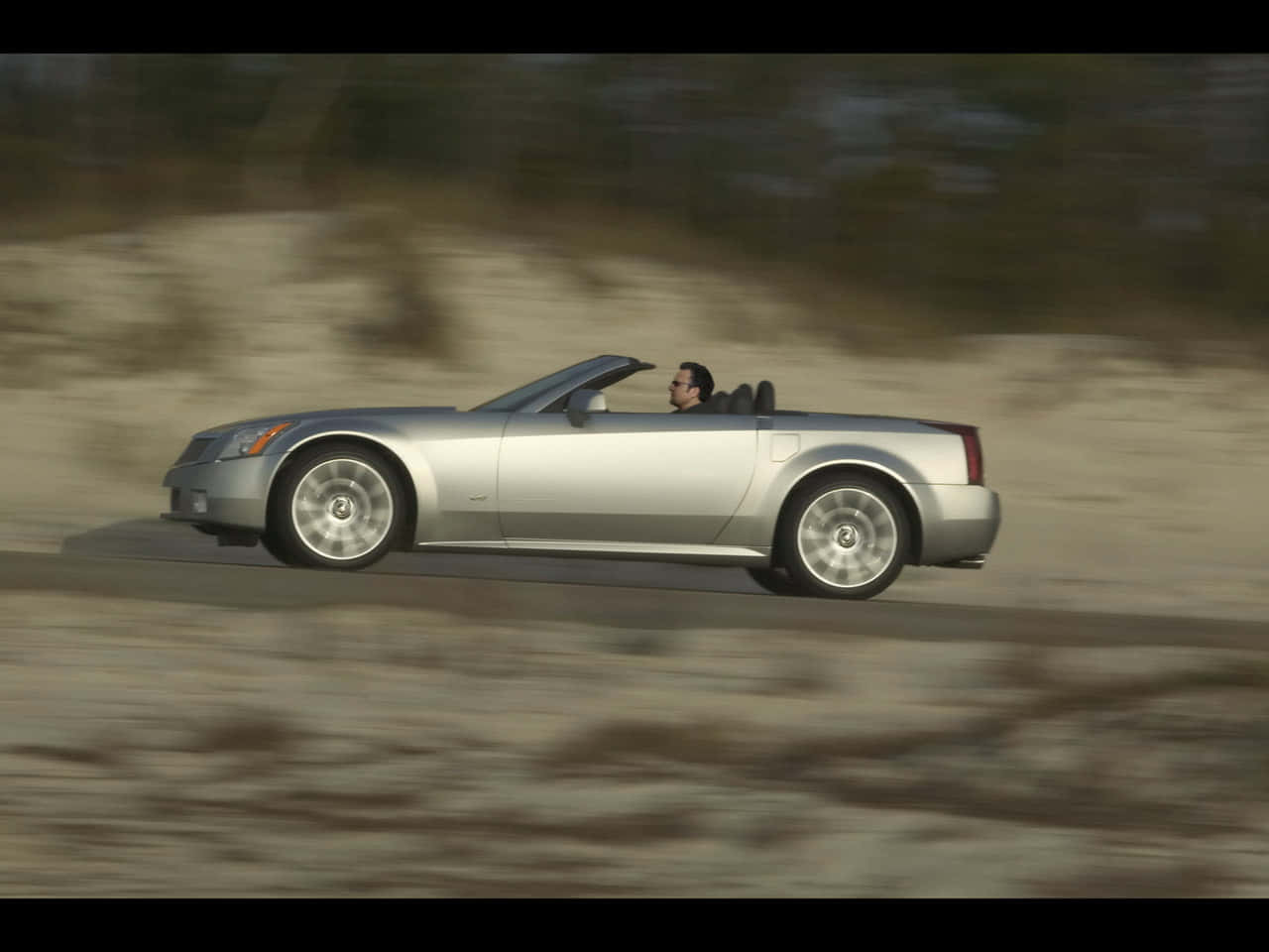 Cadillac Xlr 桌布 壁紙 — 一輛銀色敞篷跑車在沙漠公路上高速行駛，模糊的環境傳達出動感。