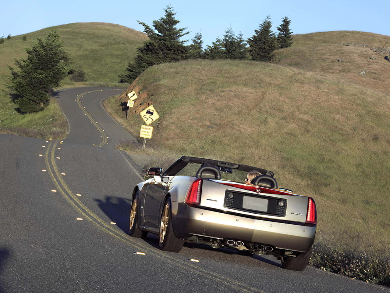 Cadillac Xlr 桌布 壁紙 — 一輛銀色敞篷跑車沿著蜿蜒的山路行駛，兩旁是長滿青草的山丘和常綠樹木，天空清澈湛藍。