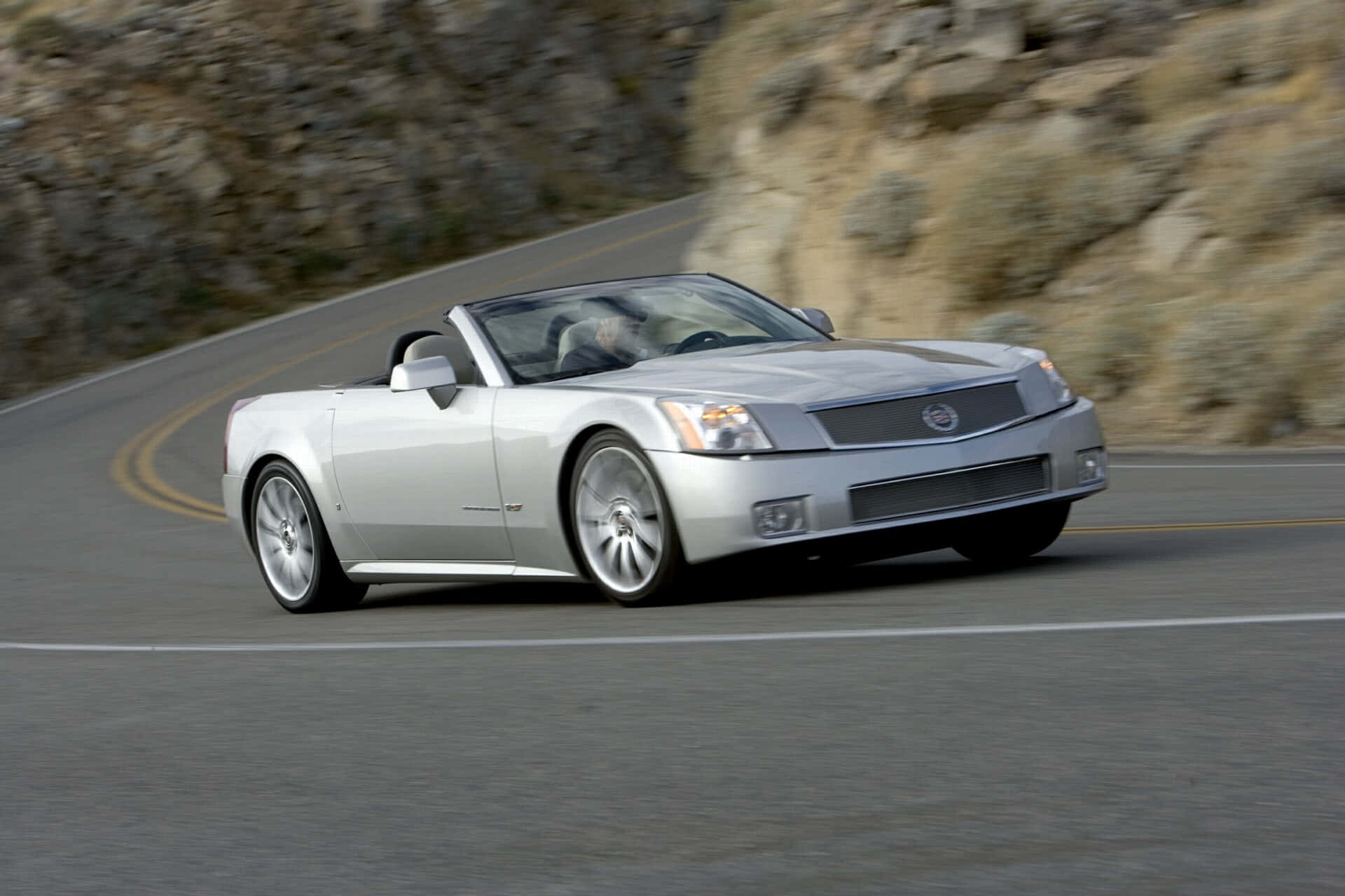 Cadillac Xlr 桌布 壁紙 — 一輛銀色敞篷跑車沿著蜿蜒的山路飛馳，背景是模糊的岩石懸崖。
