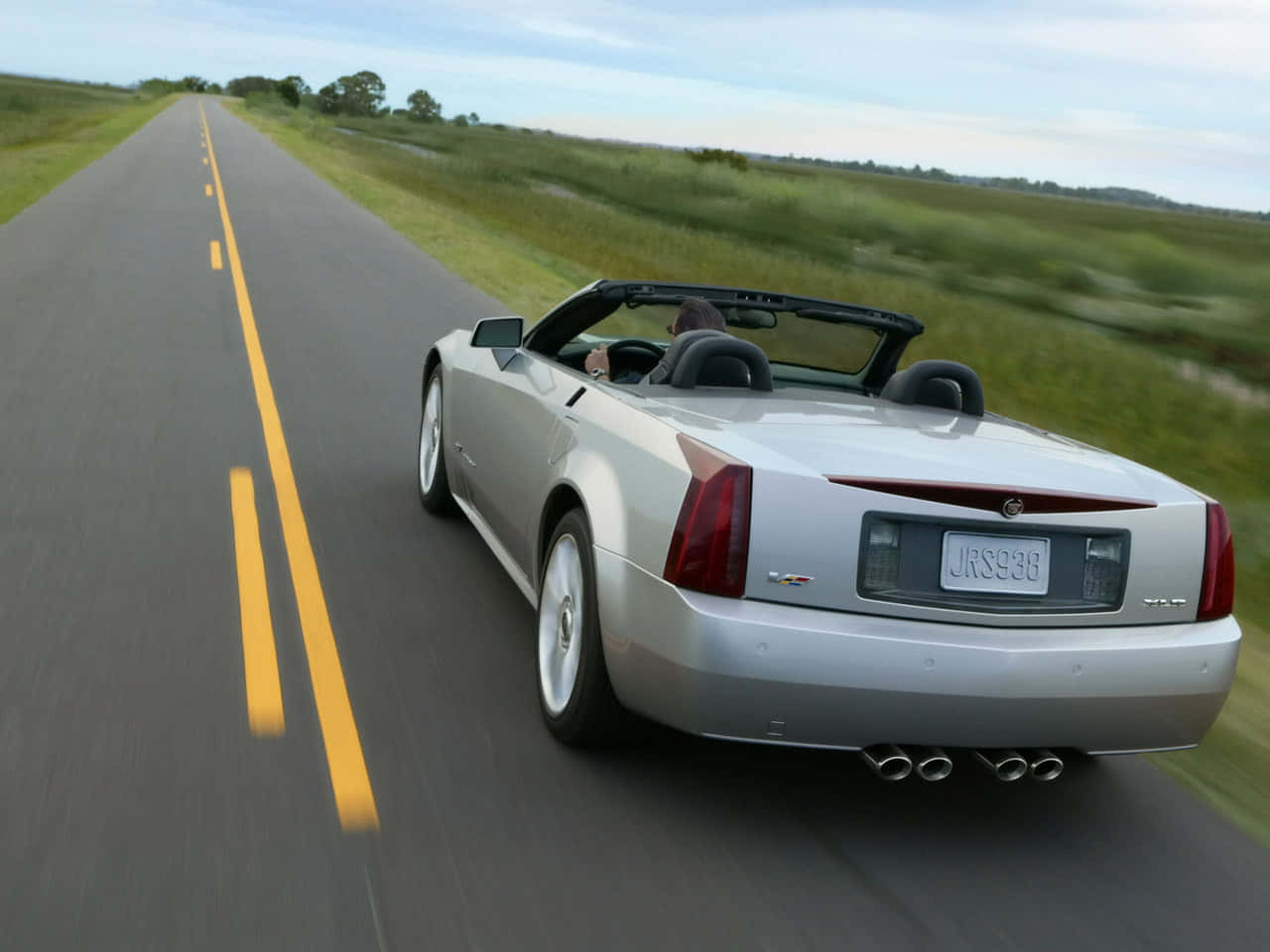 Cadillac Xlr 桌布 壁紙 — 一輛銀色敞篷車在明亮的天空下，沿著一條筆直的鄉村道路飛馳，兩旁是模糊的綠色田野和黃色標線。