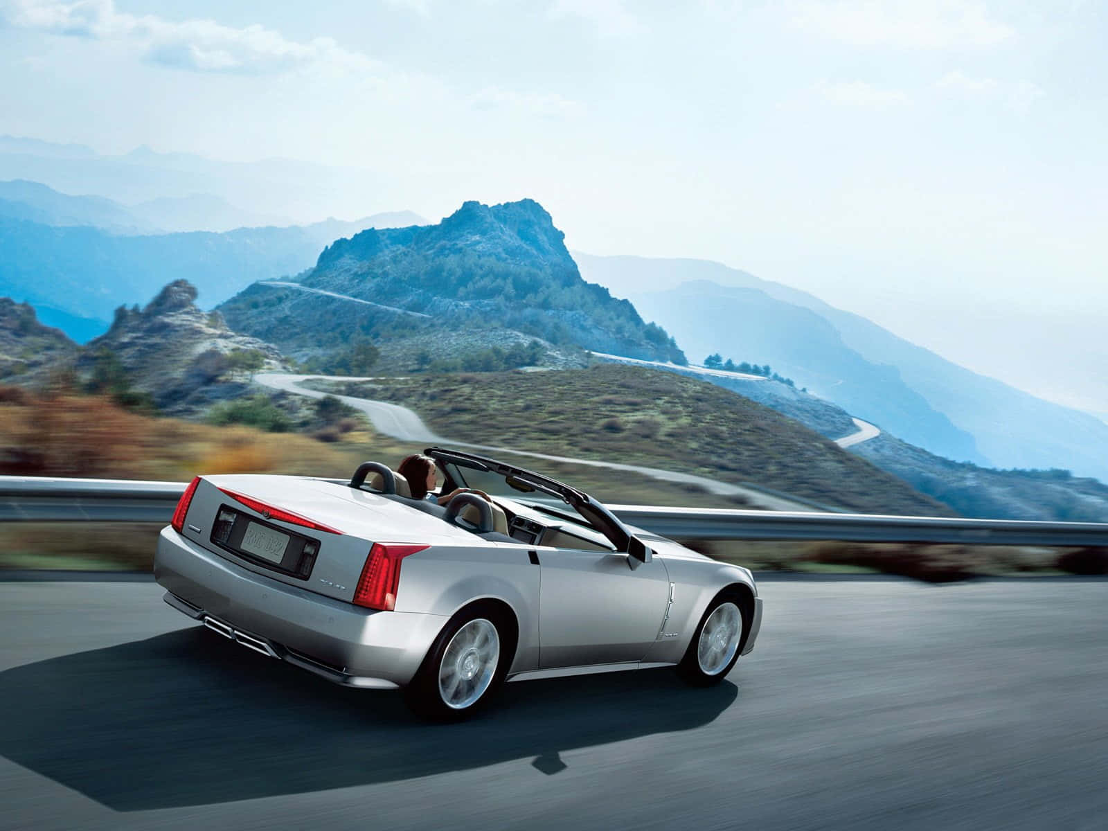 Cadillac Xlr 桌布 壁紙 — 一輛銀色敞篷車在晴朗天空下，沿著蜿蜒的山路疾駛，風景模糊，遠峰隱約可見。