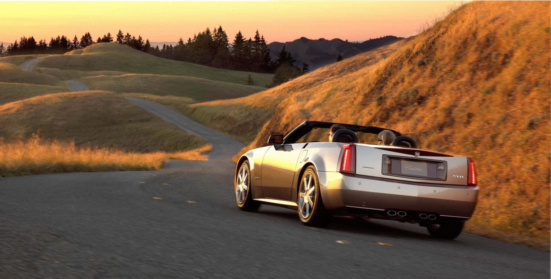 Cadillac Xlr 桌布 壁紙 — 一輛銀色敞篷車沿著蜿蜒的鄉間道路行駛，背景是金色夕陽下的起伏山丘和樹木。