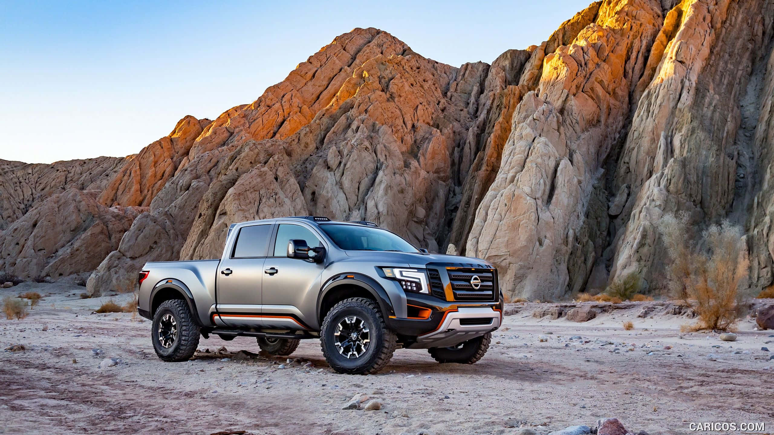 Nissan Titan 桌布 壁紙 — 一輛銀色日產Frontier皮卡車矗立在崎嶇的沙漠地形上，戲劇性的岩石懸崖沐浴在金黃色的日落光芒中。