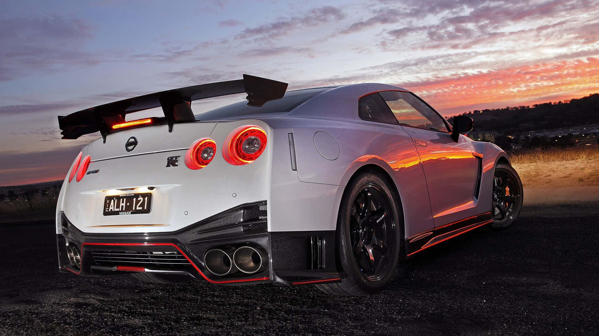 桌布 壁紙 — 一輛銀色日產GT-R R35跑車，紅色尾燈發光，停在夕陽下的礫石上。