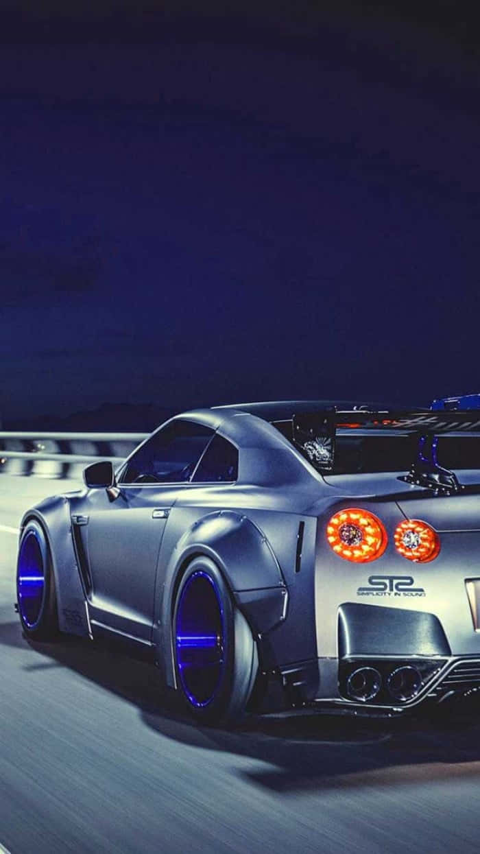 JDM 桌布 壁紙 — 一輛銀色日產GT-R，帶有發光的橙色尾燈和藍色輪圈燈，在夜間沿著黑暗的高速公路飛馳。