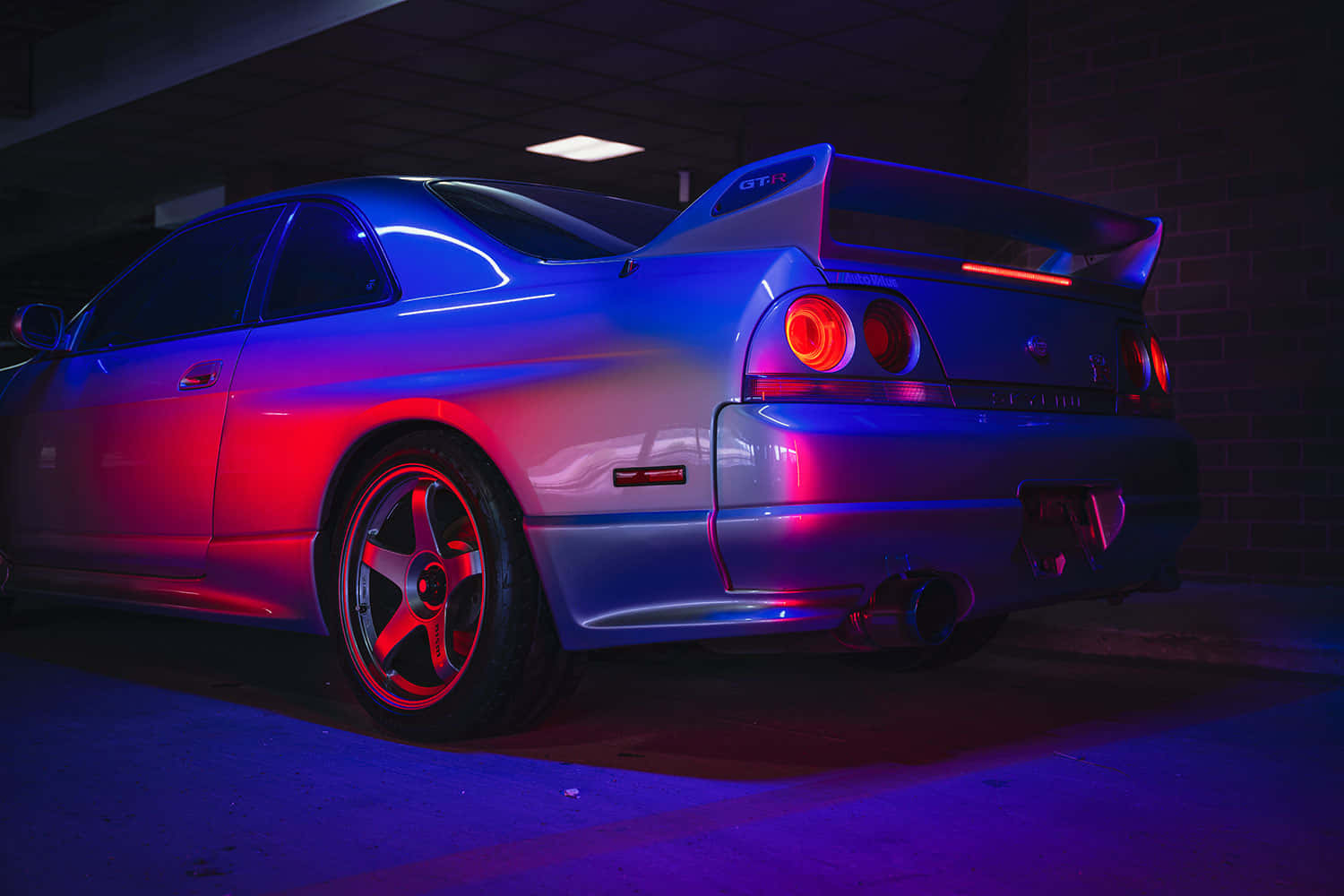 R33 GT-R 桌布 壁紙 — 一輛銀色日產GT-R跑車在昏暗的地下停車場中，在鮮豔的紅色和藍色霓虹燈下閃閃發光。