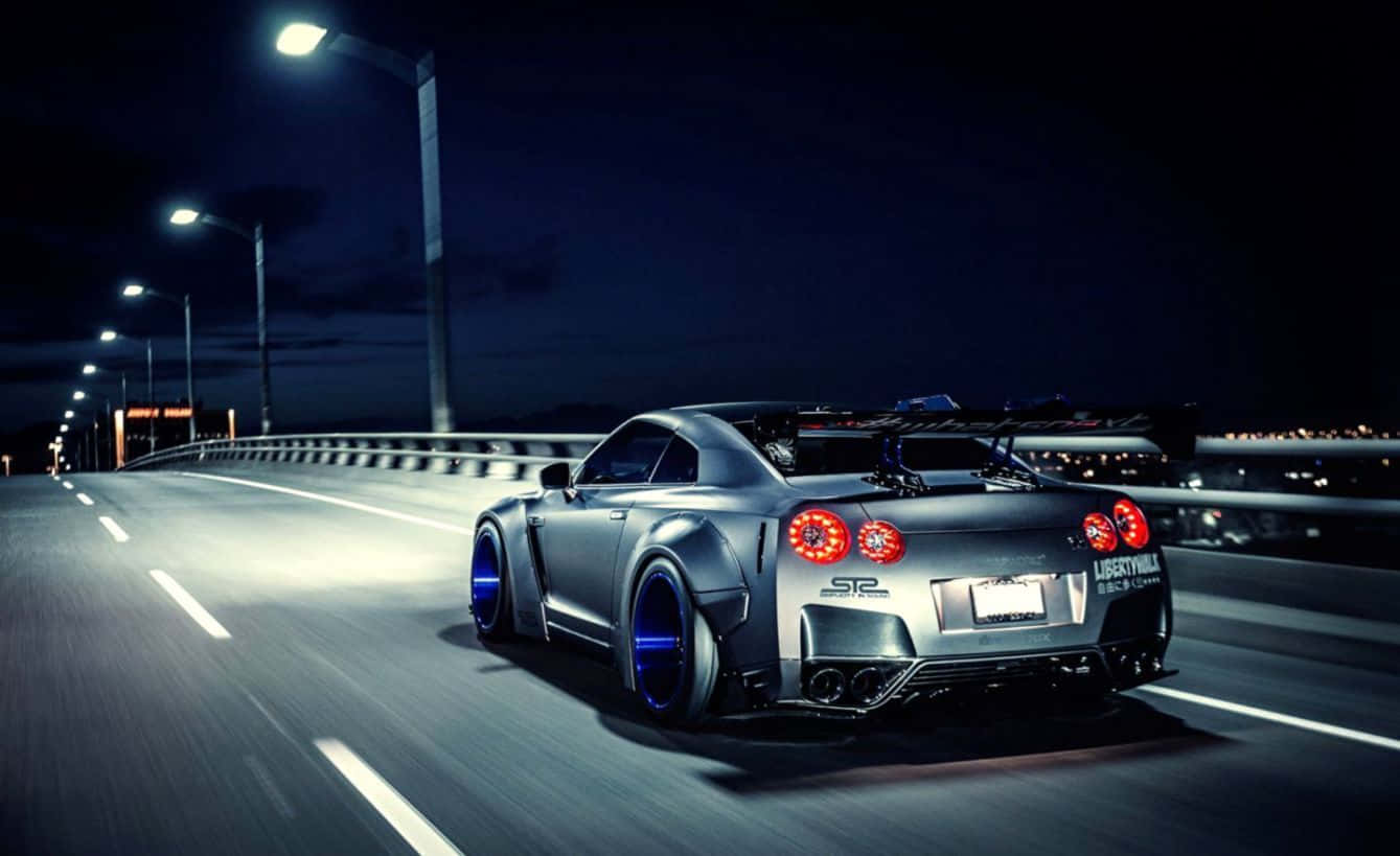 汽車 4K 桌布 壁紙 — 一輛銀色日產GT-R配藍色輪圈，在夜間有燈光的橋上以動態模糊效果疾馳。