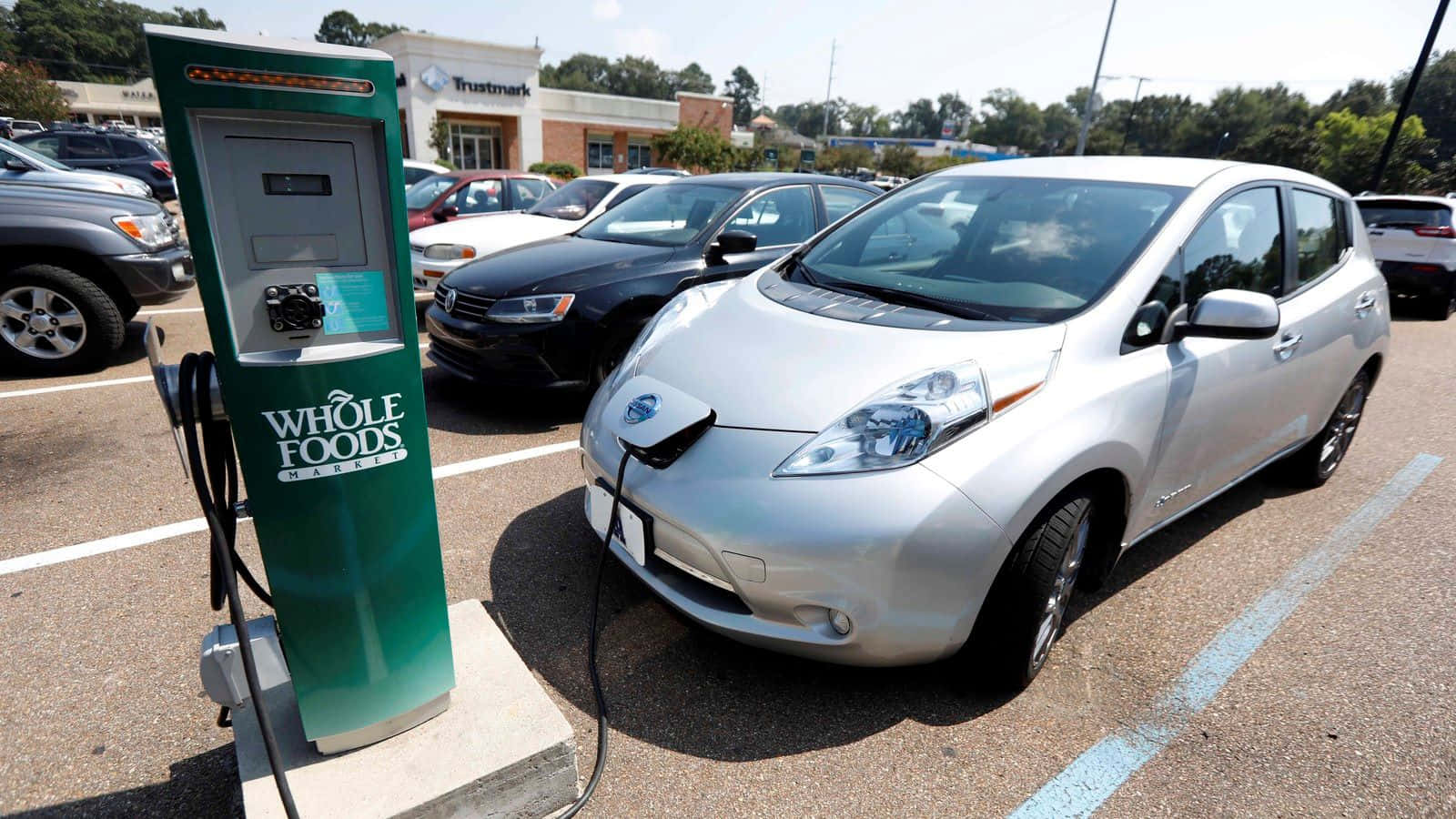 Electric Vehicle 桌布 壁紙 — 一輛銀色日產Leaf電動車在陽光明媚的停車場插入綠色Whole Foods Market充電站。