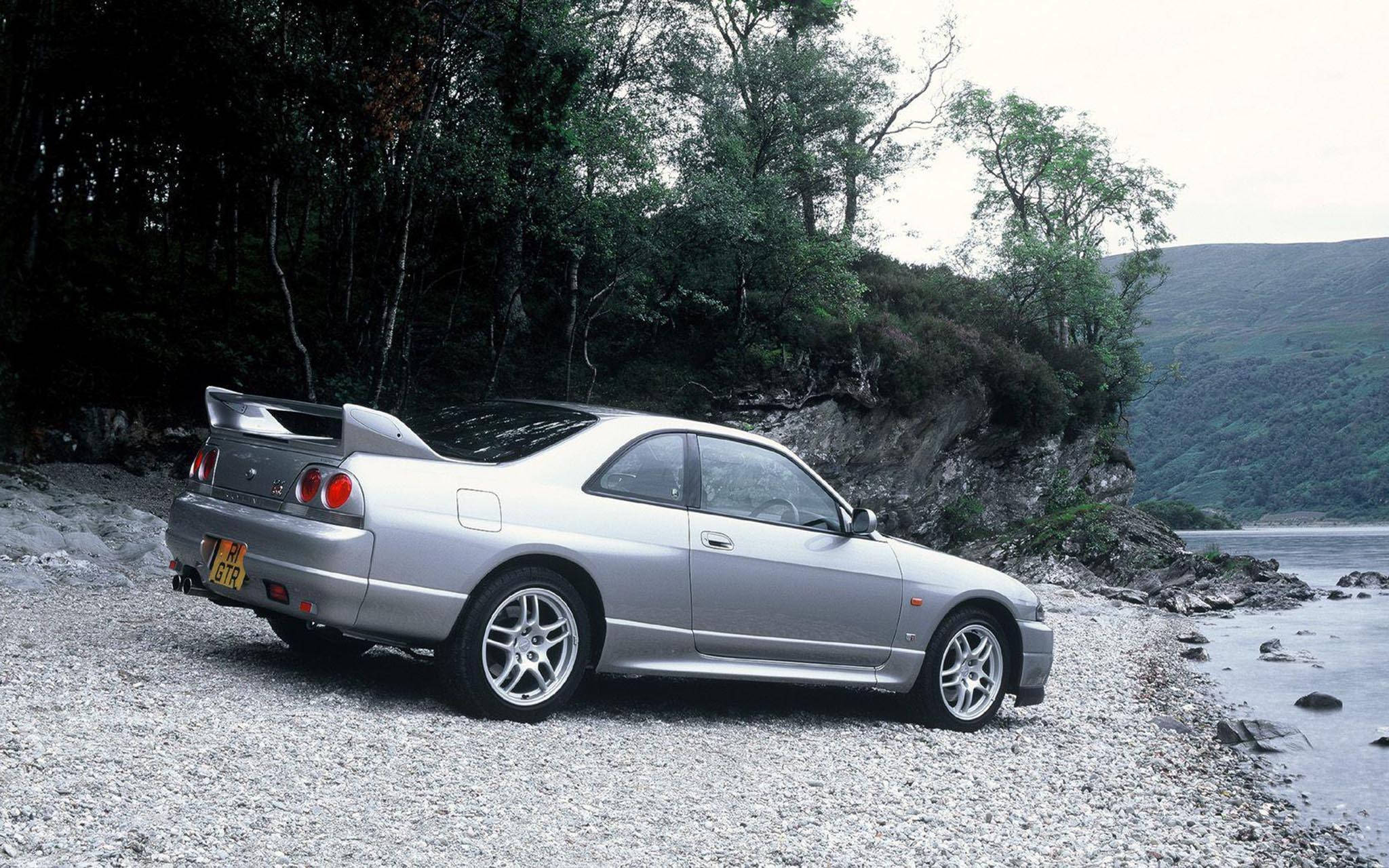 Gtr 桌布 壁紙 — 一輛銀色日產Skyline GTR停泊在平靜湖泊旁的礫石岸邊，背景是樹木繁茂的山丘。