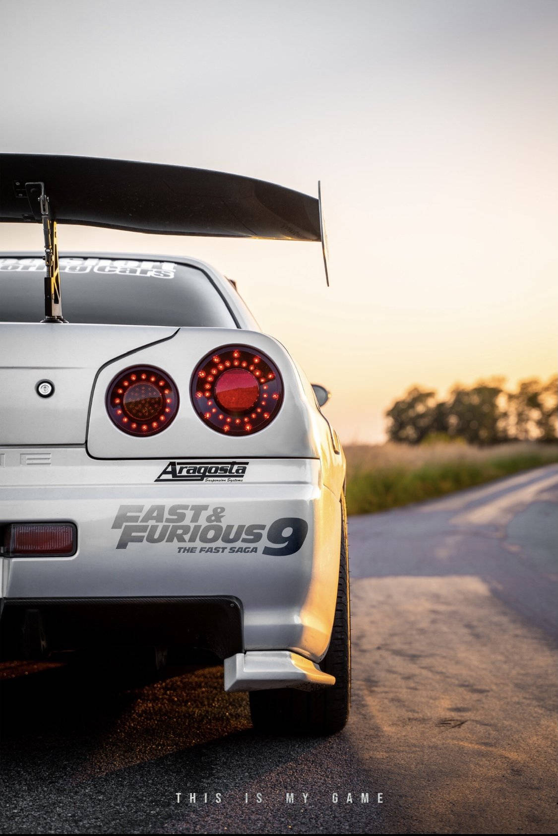 Fast And Furious Cars 桌布 壁紙 — 一輛銀色日產Skyline GT-R，帶有大型尾翼和發光的紅色尾燈，停在日落時分的鄉村道路上。