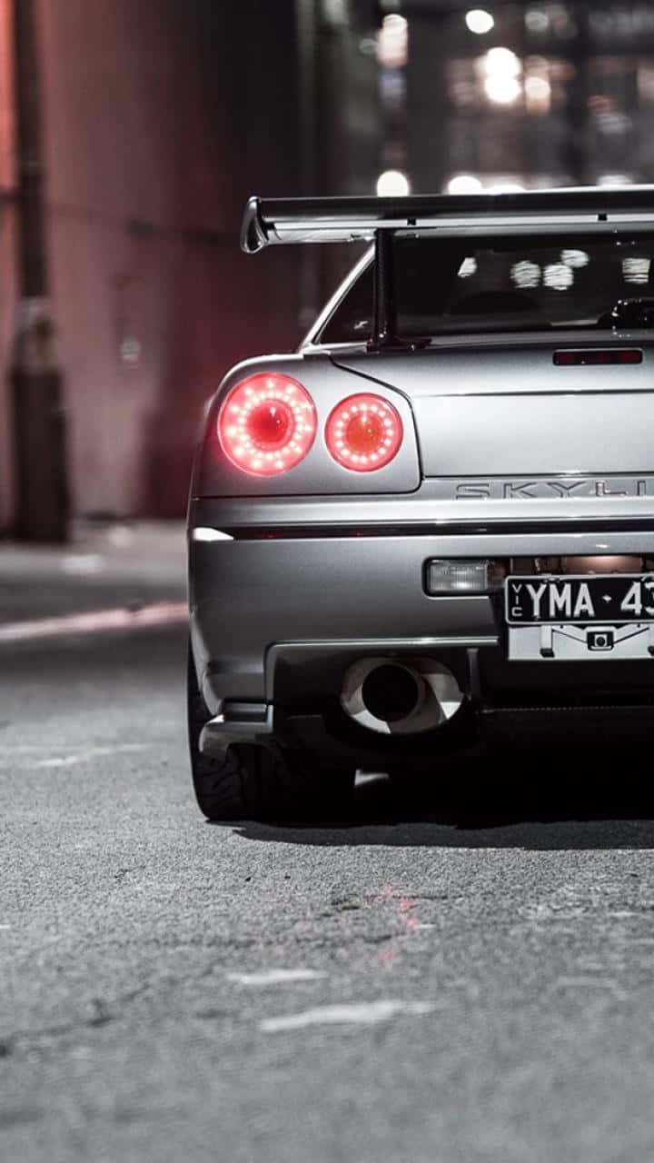 JDM 桌布 壁紙 — 一輛銀色日產Skyline GT-R，帶有發光的紅色圓形尾燈，停泊在夜晚潮濕的城市街道上。