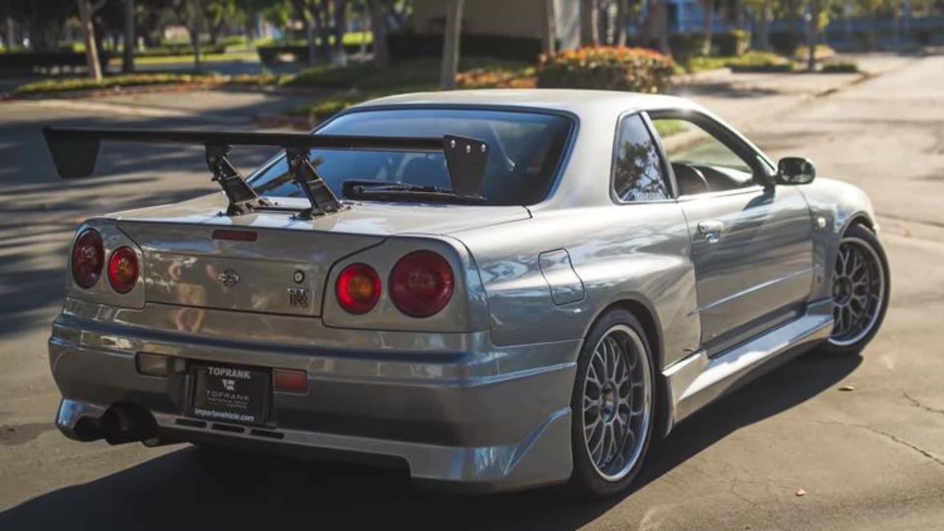 Paul Walker Skyline 桌布 壁紙 — 一輛銀色日產Skyline R34跑車，帶有大型尾翼，停泊在陽光普照的郊區街道上。