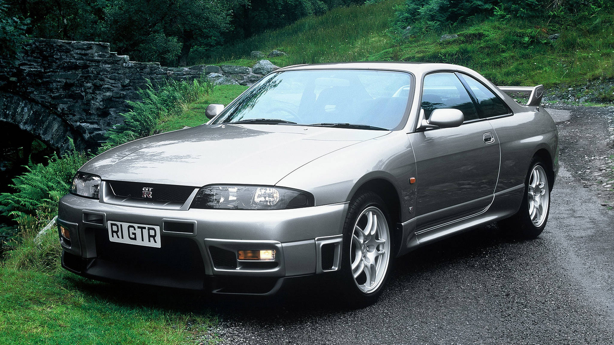 Gtr 桌布 壁紙 — 一輛銀色日產Skyline R34 GTR停放在茂密綠色森林中石牆旁的濕碎石路上。
