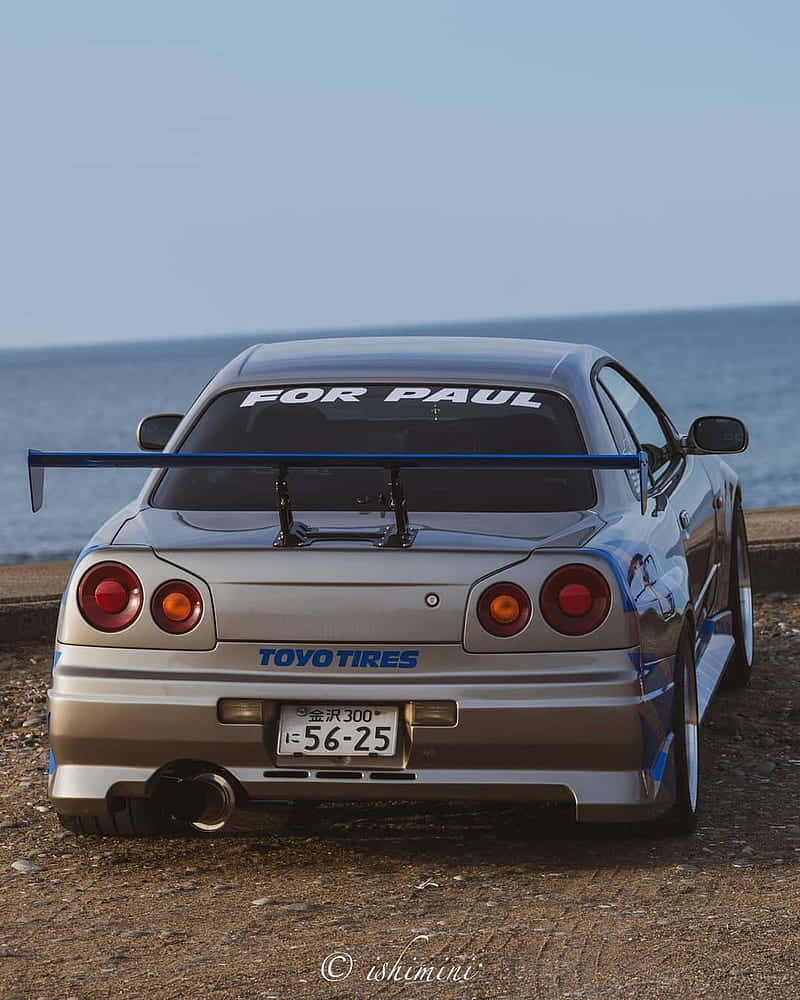 Paul Walker Skyline 桌布 壁紙 — 一輛銀色日產Skyline R34，帶有藍色尾翼和「FOR PAUL」貼紙，停泊在俯瞰海洋的岩石海岸上。