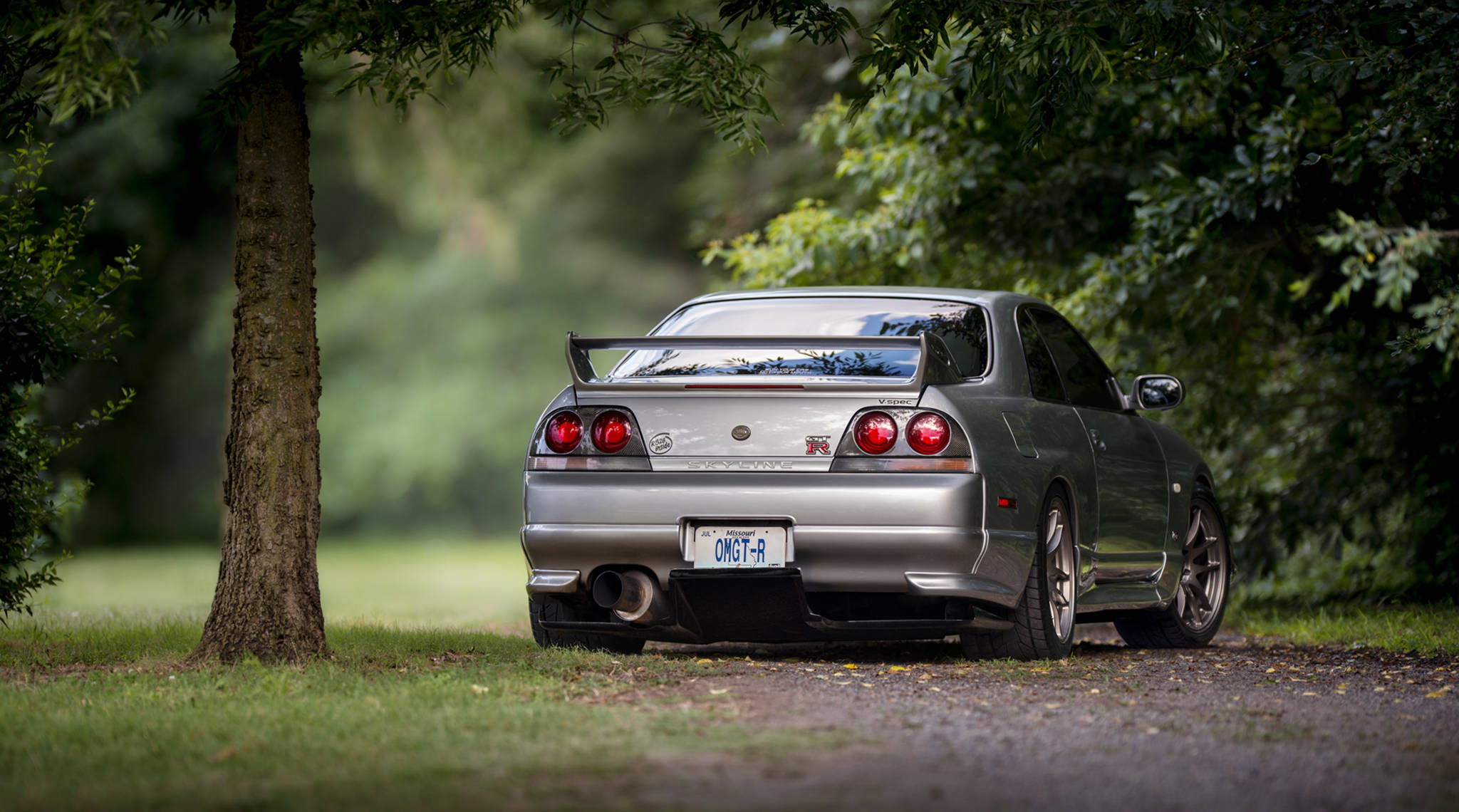 Gtr 桌布 壁紙 — 一輛銀色日產Skyline R34 GT-R停放在碎石路上，周圍是綠樹成蔭的寧靜森林環境。