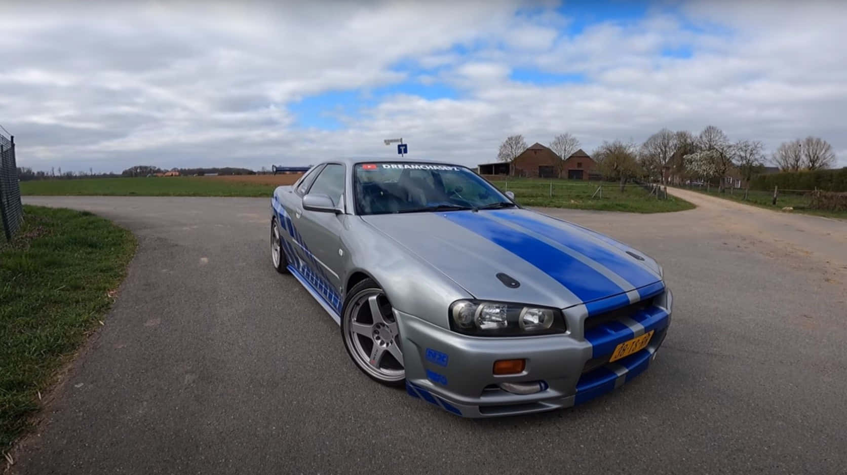 Paul Walker Skyline 桌布 壁紙 — 一輛銀色日產Skyline R34，帶有醒目的藍色賽車條紋，停在鄉村瀝青路上，天空部分多雲。