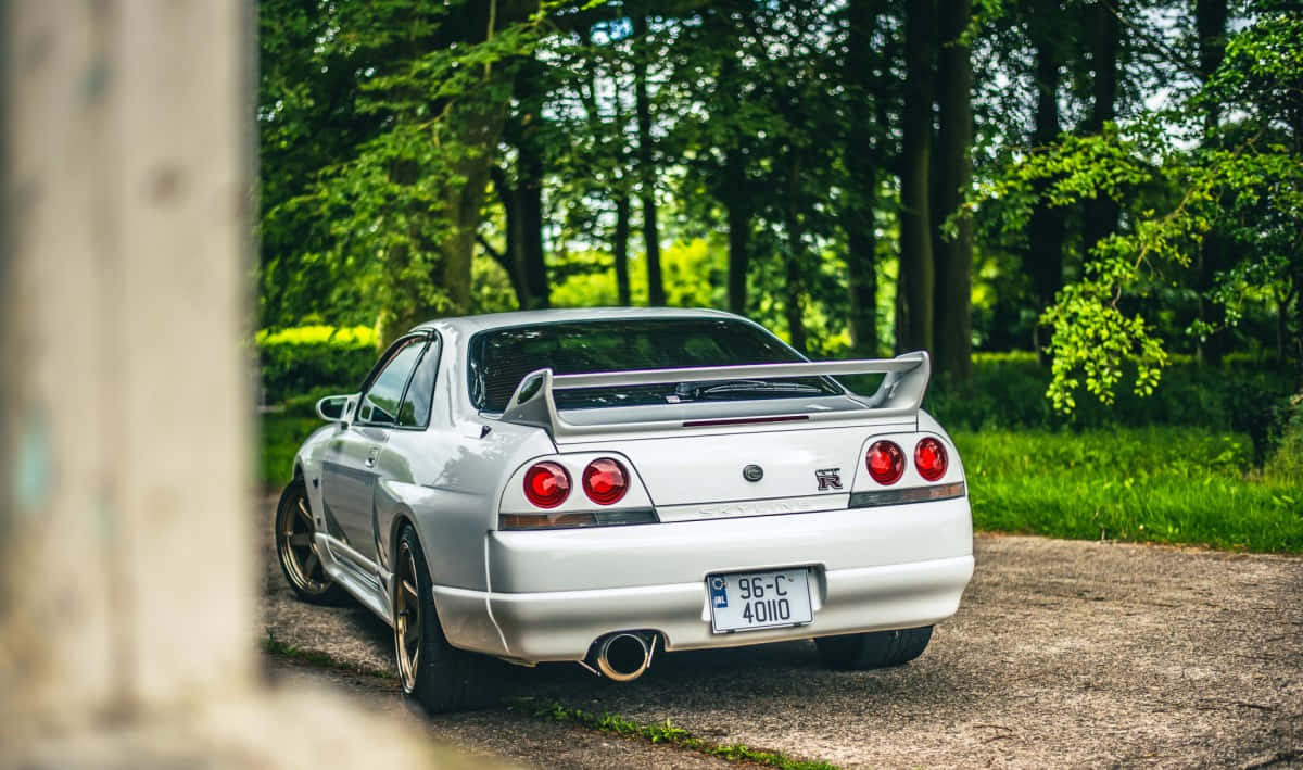 R33 GT-R 桌布 壁紙 — 一輛銀色日產Skyline R33 GT-R停放在碎石路上，周圍是茂密的綠樹，在明亮的日光下。