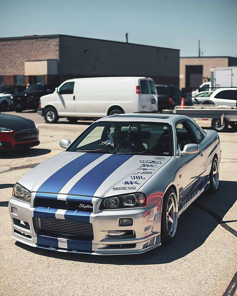 Paul Walker Skyline 桌布 壁紙 — 一輛銀色日產Skyline R34，帶有醒目的藍色賽車條紋，停放在陽光明媚的瀝青停車場，周圍有其他車輛。