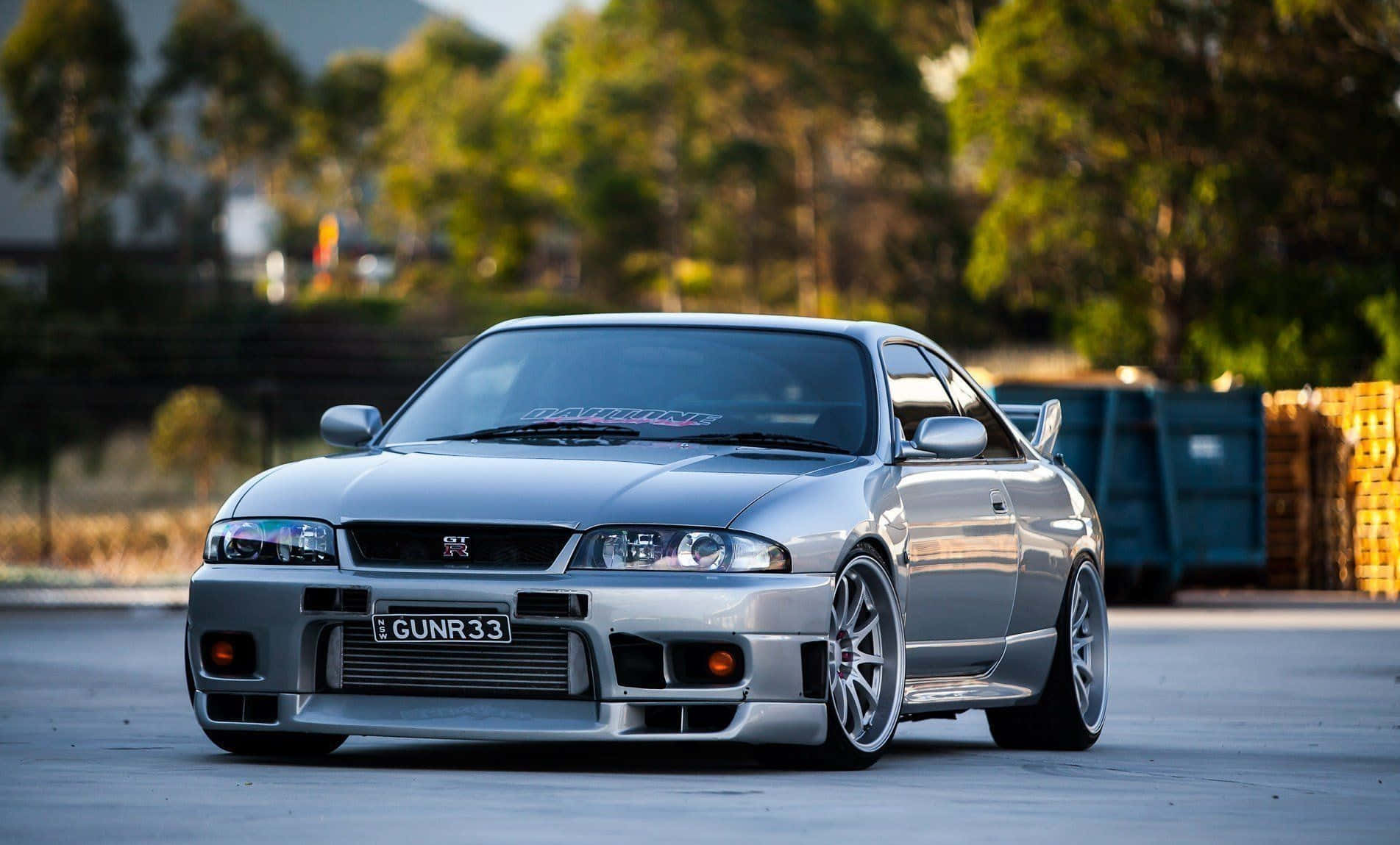 R33 GT-R 桌布 壁紙 — 一輛銀色日產Skyline R34跑車，配備訂製輪圈及前保險桿，停放在一個被樹木和垃圾箱環繞的混凝土場地上。