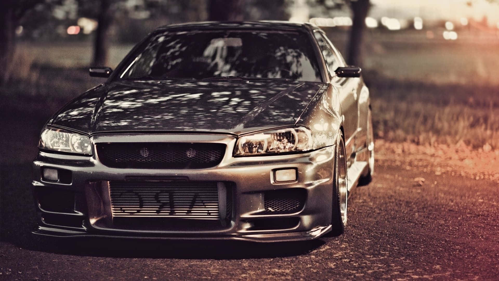 桌布 壁紙 — 一輛銀色日產Skyline R34飄移車停泊在碎石路上，正值黃金時段，背景有樹木和模糊的城市燈光。