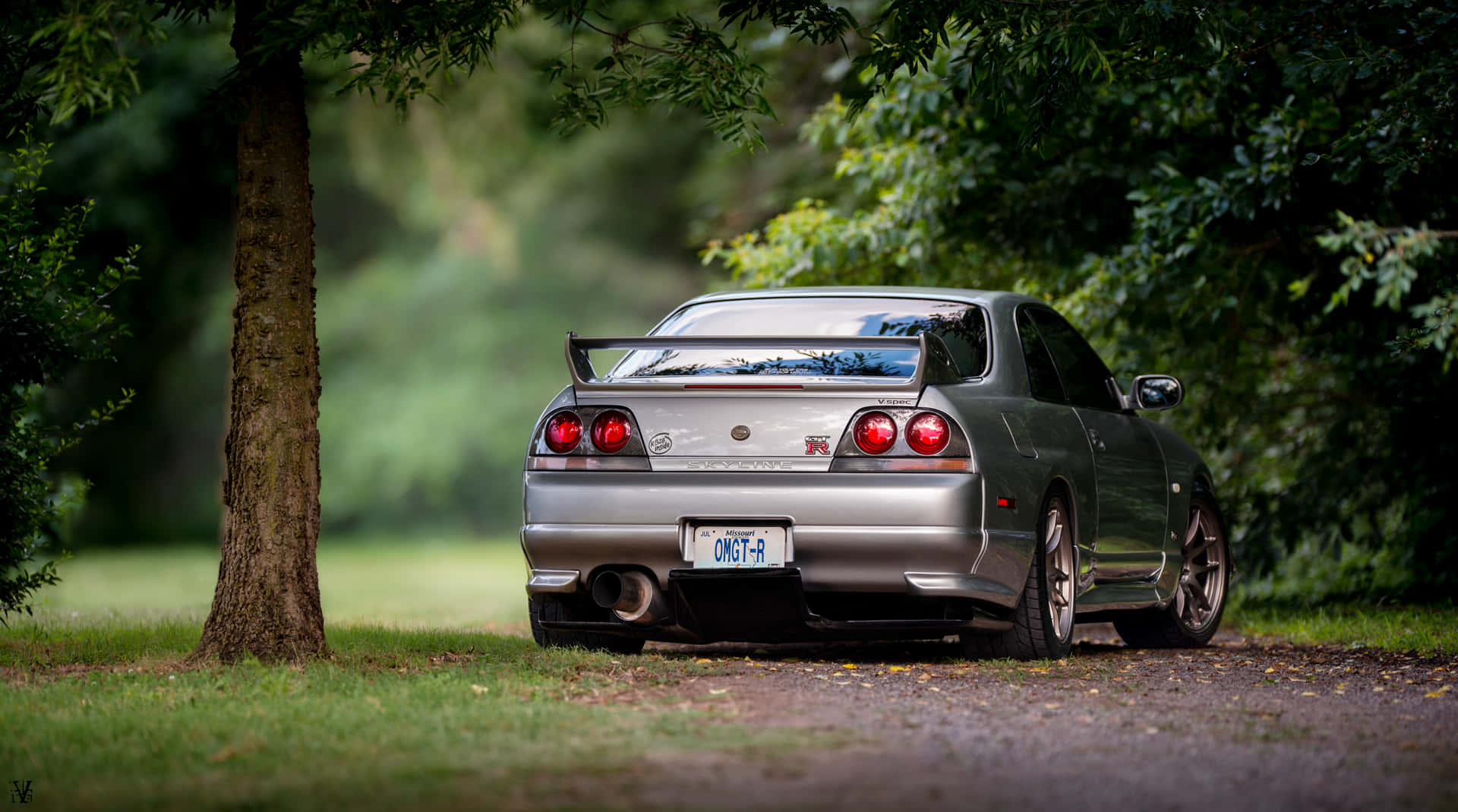 R33 GT-R 桌布 壁紙 — 一輛銀色日產Skyline R34停放在碎石小徑上，周圍是茂密的綠樹，光線柔和自然。