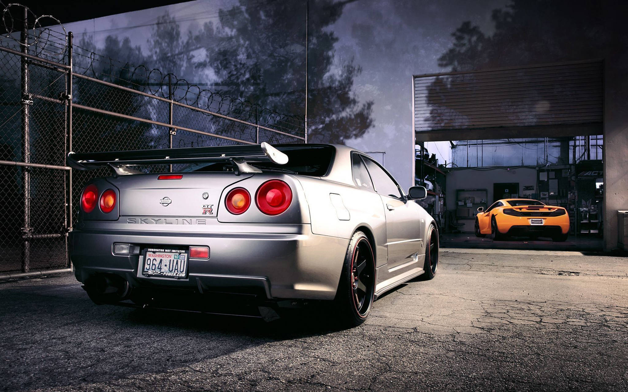 Gtr 桌布 壁紙 — 一輛銀色日產Skyline R34配紅色尾燈，停放在一個粗糙的城市車庫內，裡面可見一輛橙色跑車。