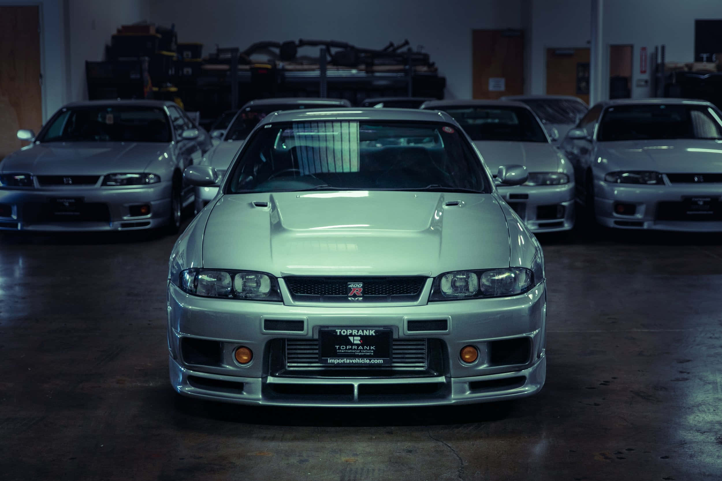 R33 GT-R 桌布 壁紙 — 一輛銀色日產Skyline R34 GT-R在昏暗的車庫中，被其他經典車包圍，面向前方。