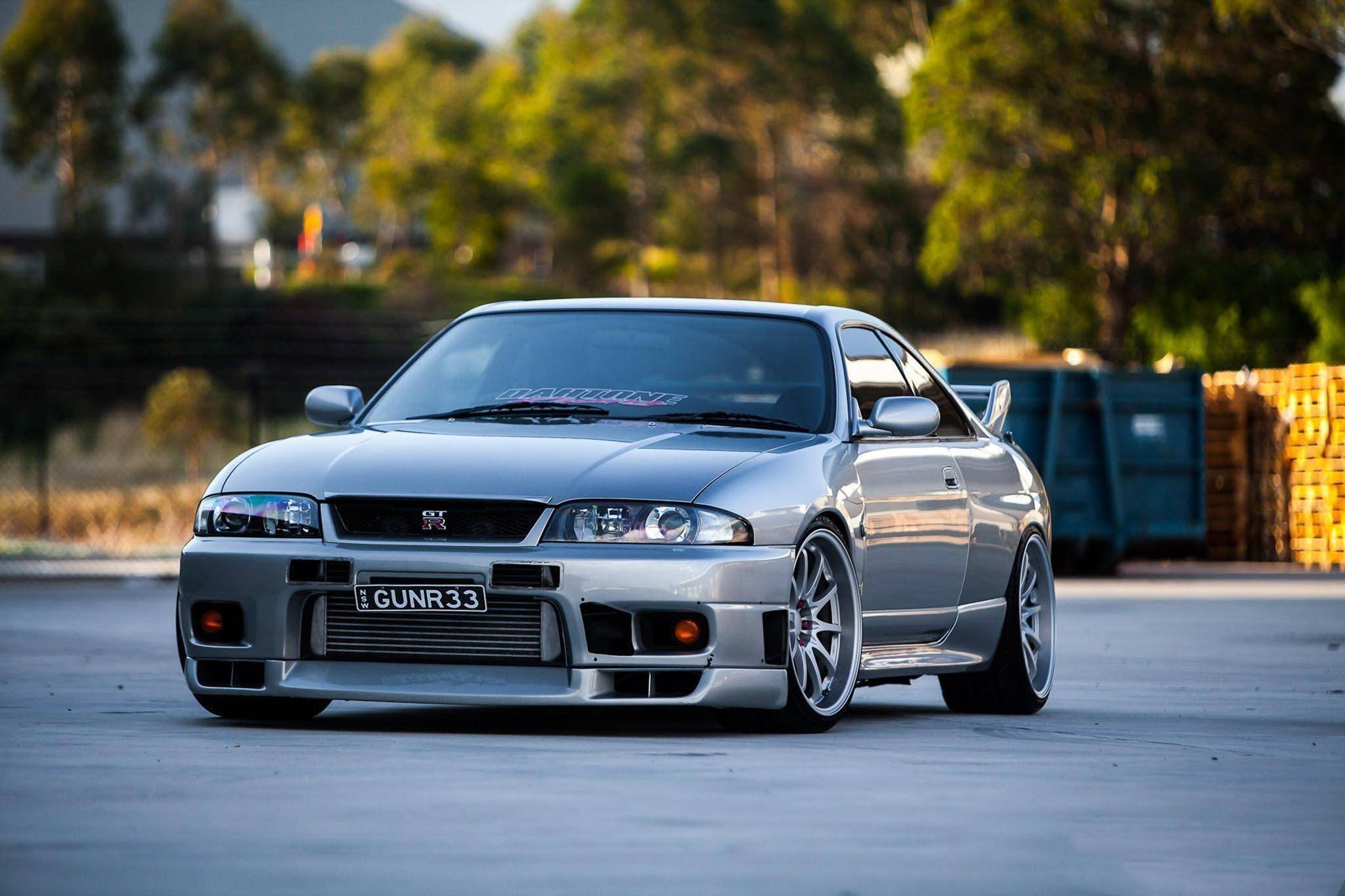 Gtr 桌布 壁紙 — 一輛銀色日產Skyline R34跑車，配備訂製輪圈和降低懸掛，停放在混凝土上，背景有樹木和垃圾箱。