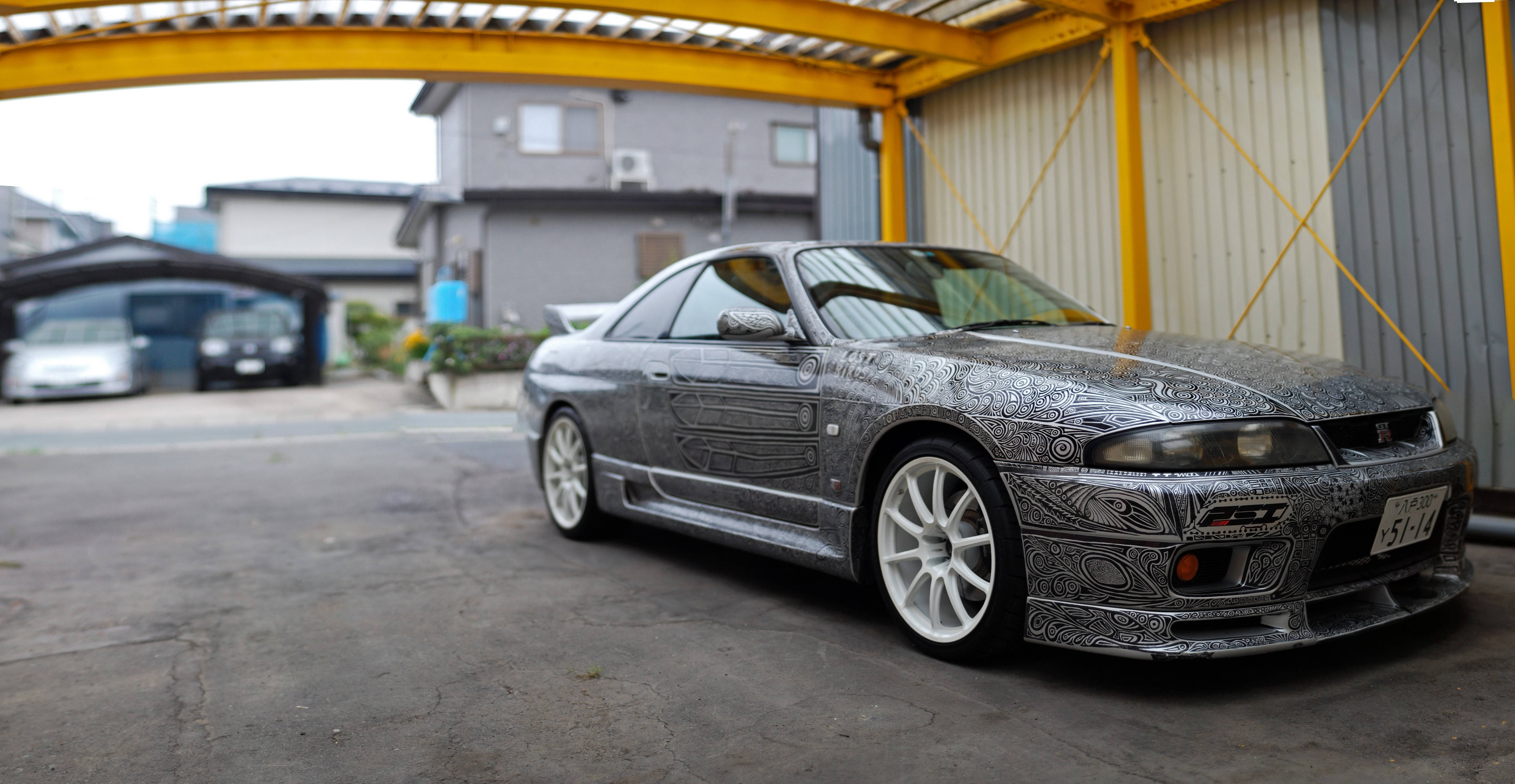 Gtr 桌布 壁紙 — 一輛銀色日產Skyline，帶有複雜的黑色部落風格貼膜，停放在住宅車道的黃色金屬車棚下。