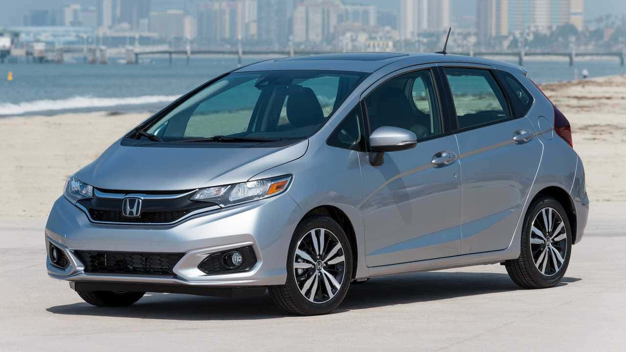 Compact Car 桌布 壁紙 — 一輛銀色本田Fit停放在陽光沙灘上，背景有城市天際線和海洋。