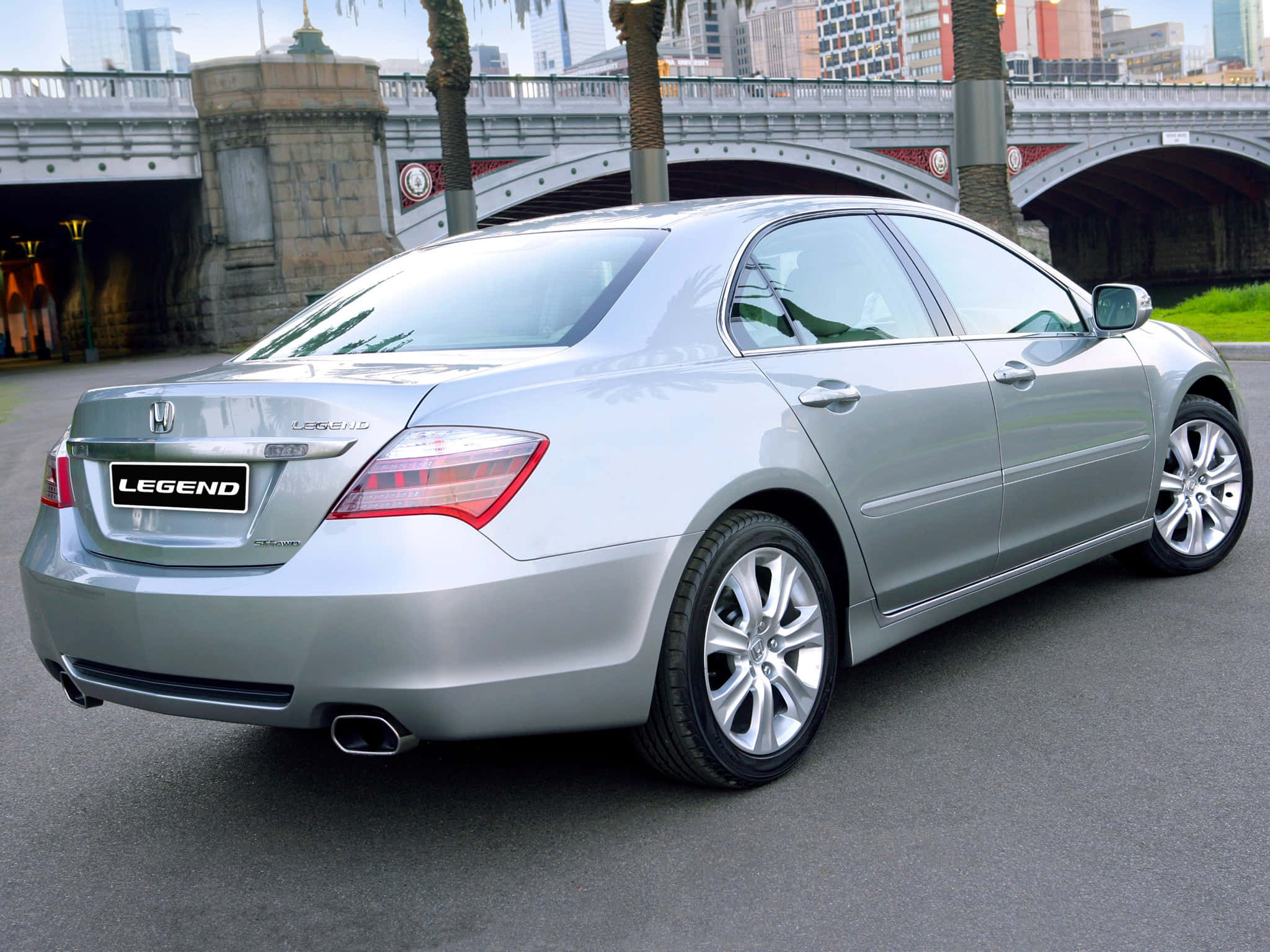 Honda Legend 桌布 壁紙 — 一輛銀色本田Legend轎車停放在拱形石橋下，背景有棕櫚樹和城市建築。