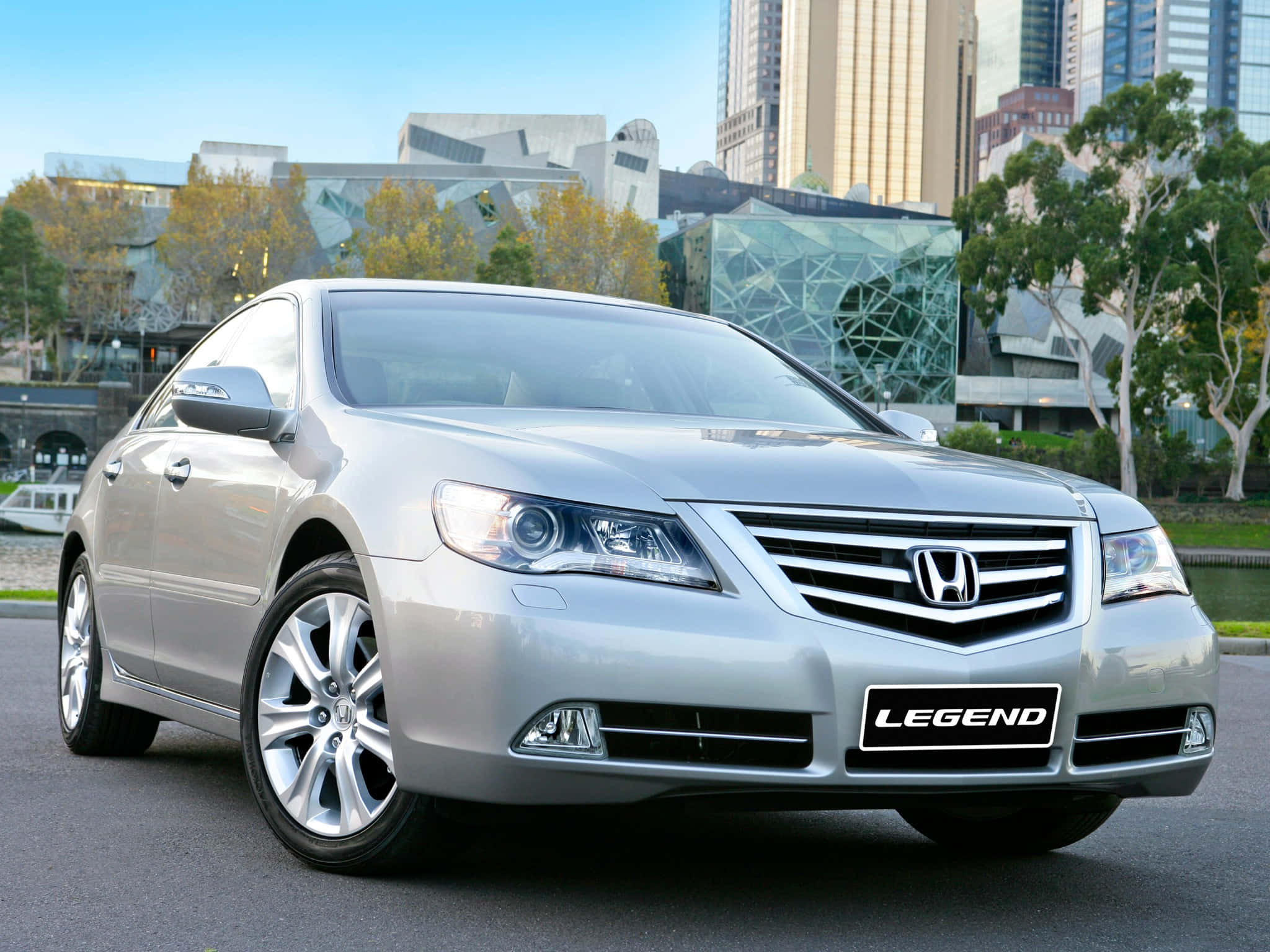 Honda Legend 桌布 壁紙 — 一輛銀色本田Legend轎車在明亮的日光下閃閃發光，停放在現代玻璃建築和秋樹附近。