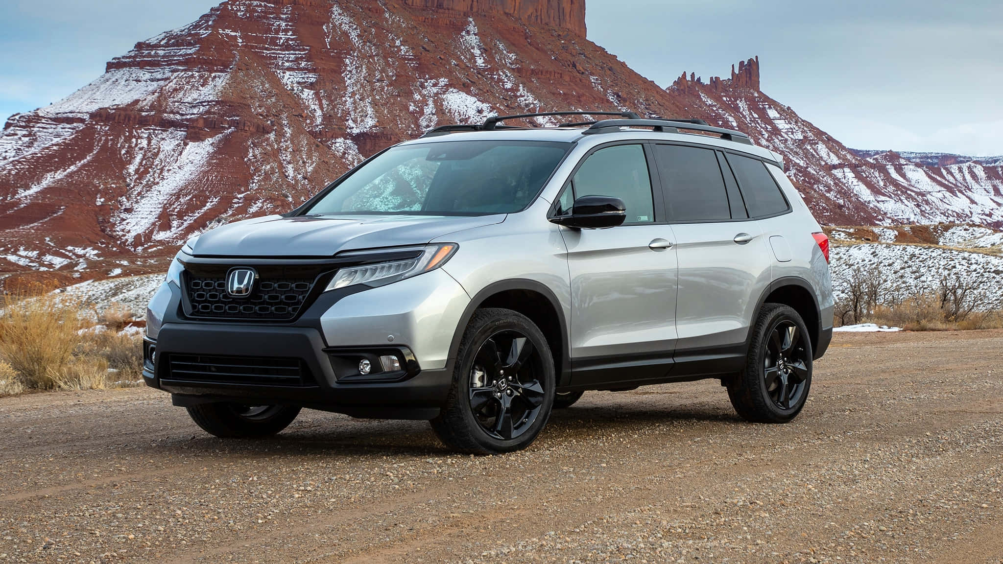 Honda Passport 桌布 壁紙 — 一輛銀色本田Pilot SUV，配黑色輪圈，停在碎石地上，前方是覆蓋薄雪的紅色岩層。