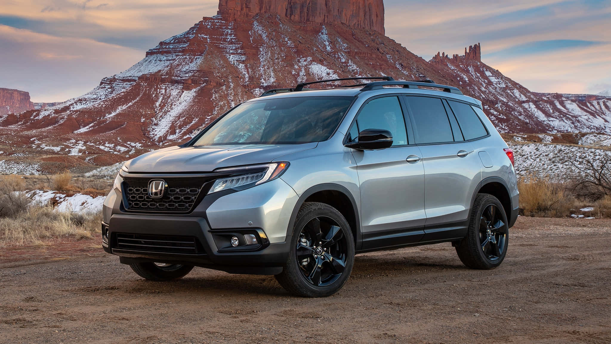 Honda Passport 桌布 壁紙 — 一輛銀色本田Pilot SUV，配黑色輪圈，停泊在日落時分被雪覆蓋的紅色岩層前的泥土路上。