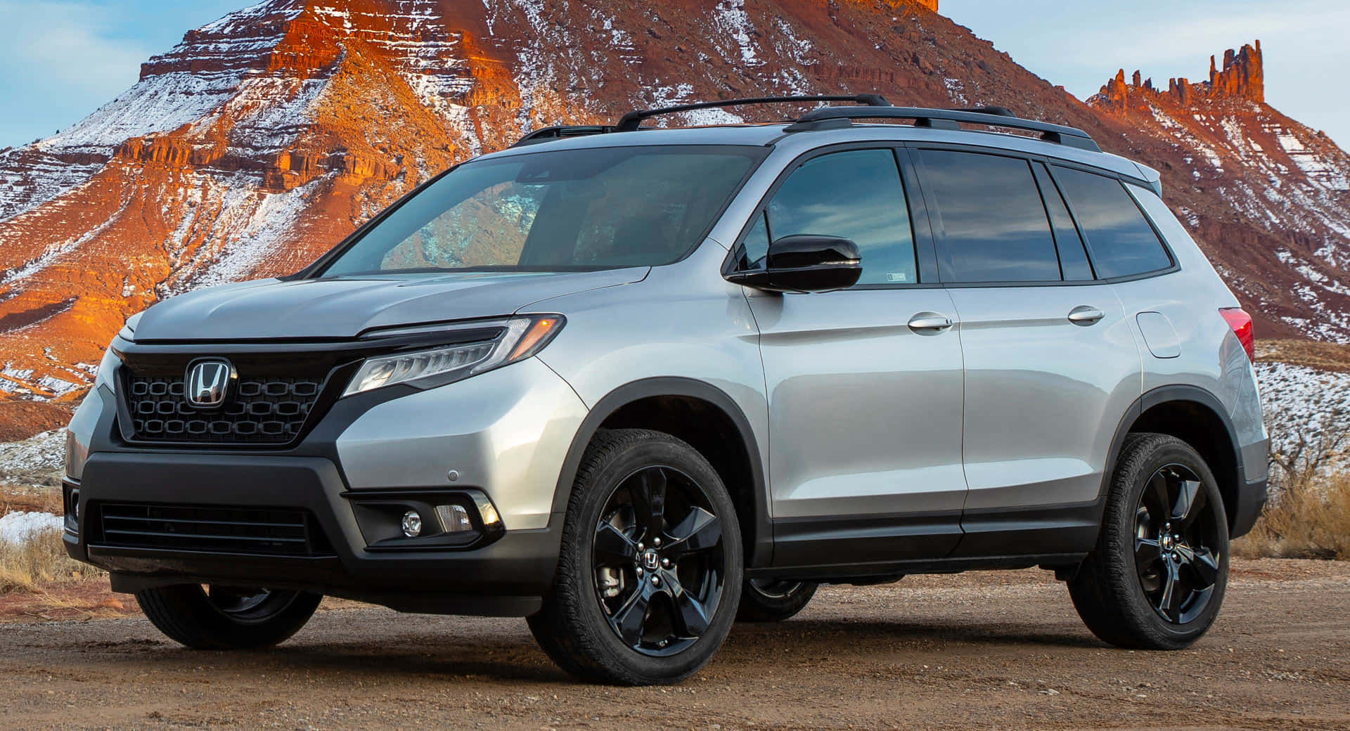 Honda Passport 桌布 壁紙 — 一輛銀色本田Pilot SUV，帶黑色飾邊，停在泥土上，背景是晴朗天空下覆蓋著薄雪的紅色岩層。