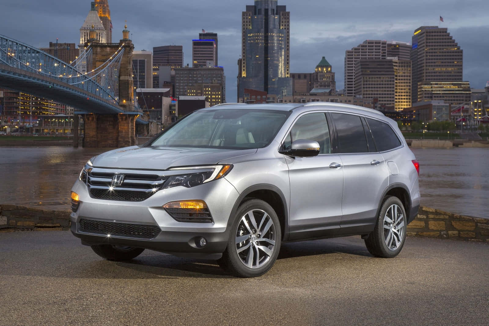 Honda Pilot 桌布 壁紙 — 一輛銀色本田Pilot SUV停泊在河邊，背景是暮色中的城市天際線和亮燈的橋樑。