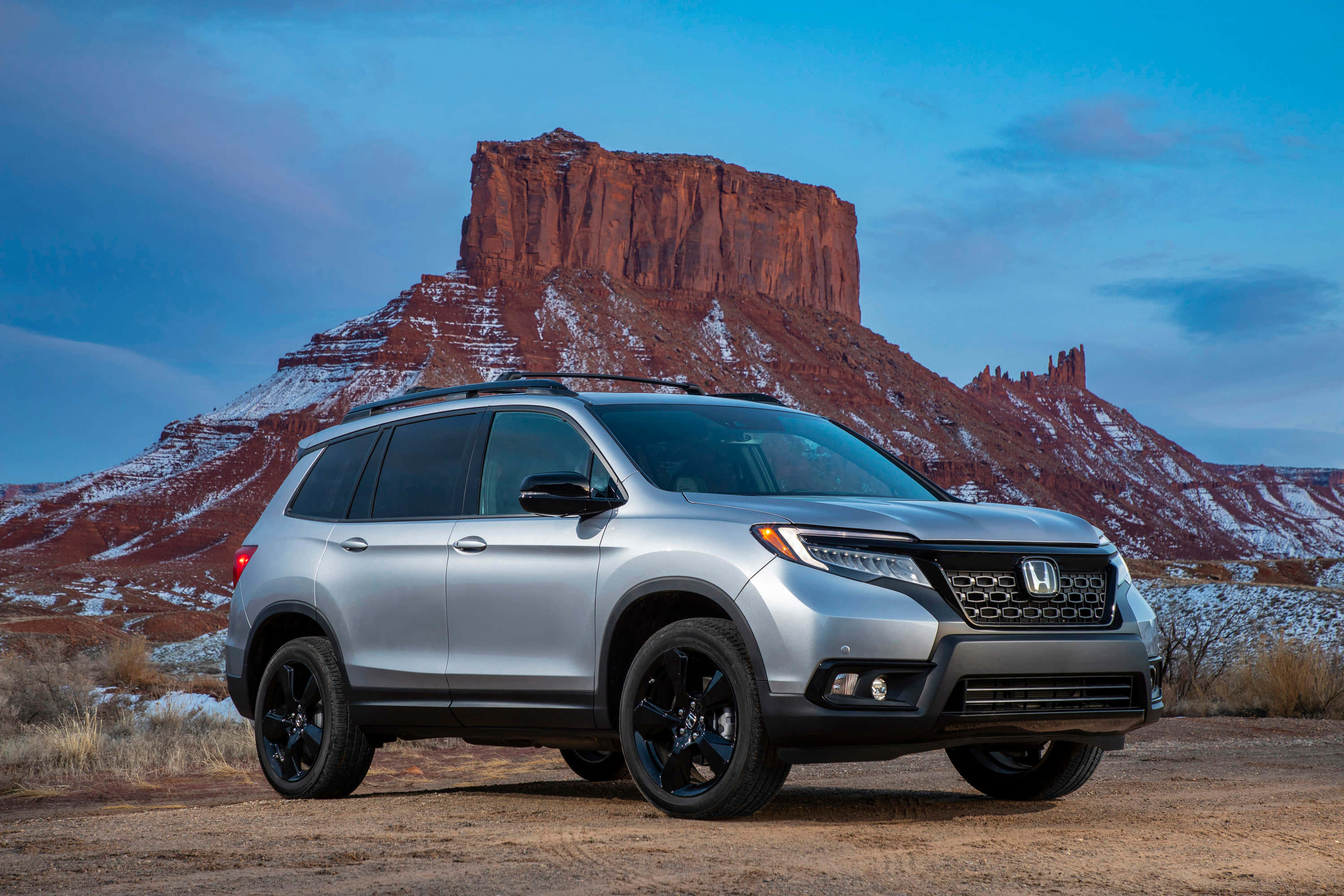 Honda Passport 桌布 壁紙 — 一輛銀色本田Pilot SUV配黑色輪圈，在暮色天空下，站在沙漠道路上，前方是被雪覆蓋的紅色岩石孤峰。