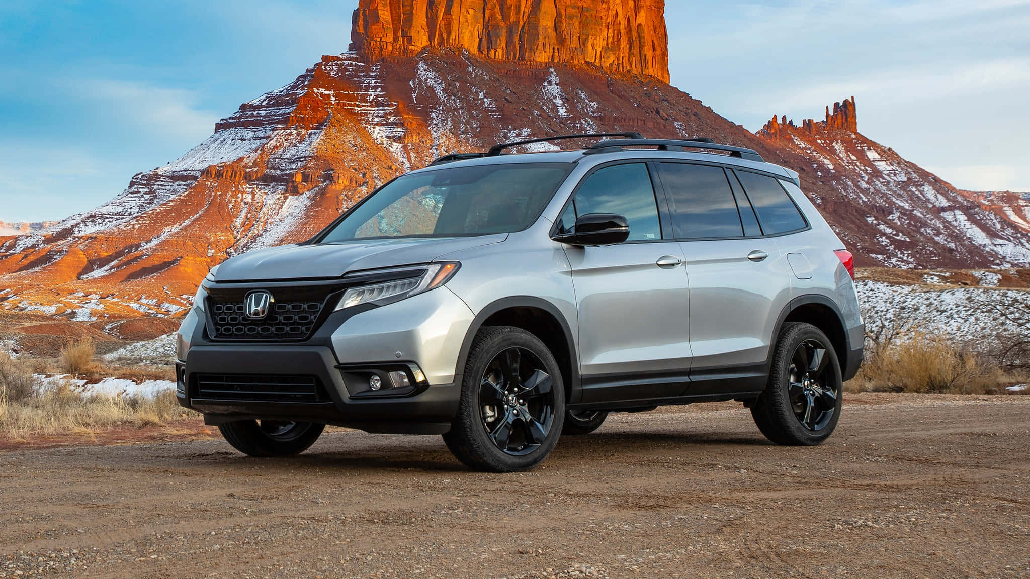Honda Passport 桌布 壁紙 — 一輛銀色本田Pilot SUV配黑色輪圈，停放在泥土上，背景是白雪覆蓋的紅色岩層，天空清澈湛藍。