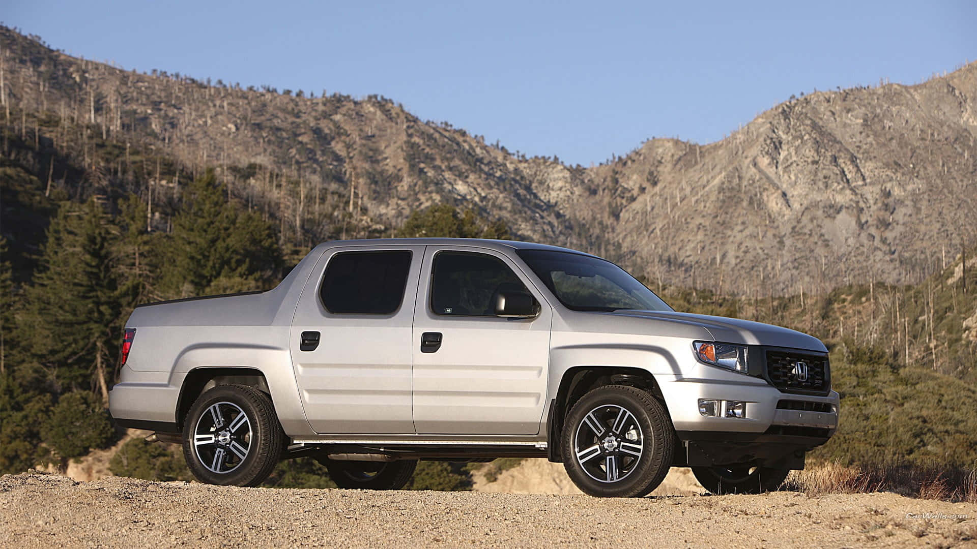 Honda Ridgeline 桌布 壁紙 — 一輛銀色本田Ridgeline皮卡車停在泥路上，背景是崎嶇的山脈和常綠樹，天空清澈湛藍。