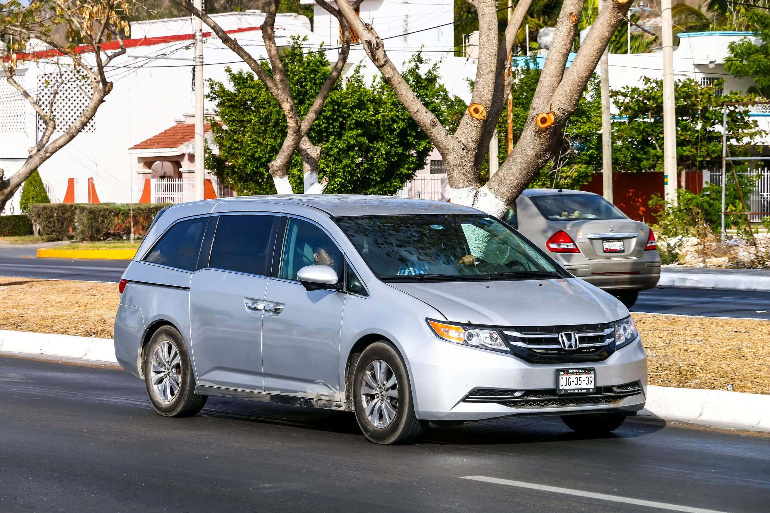 Honda Odyssey 桌布 壁紙 — 一輛銀色本田小型貨車在陽光明媚的街道上行駛，街道兩旁種滿樹木和住宅建築。