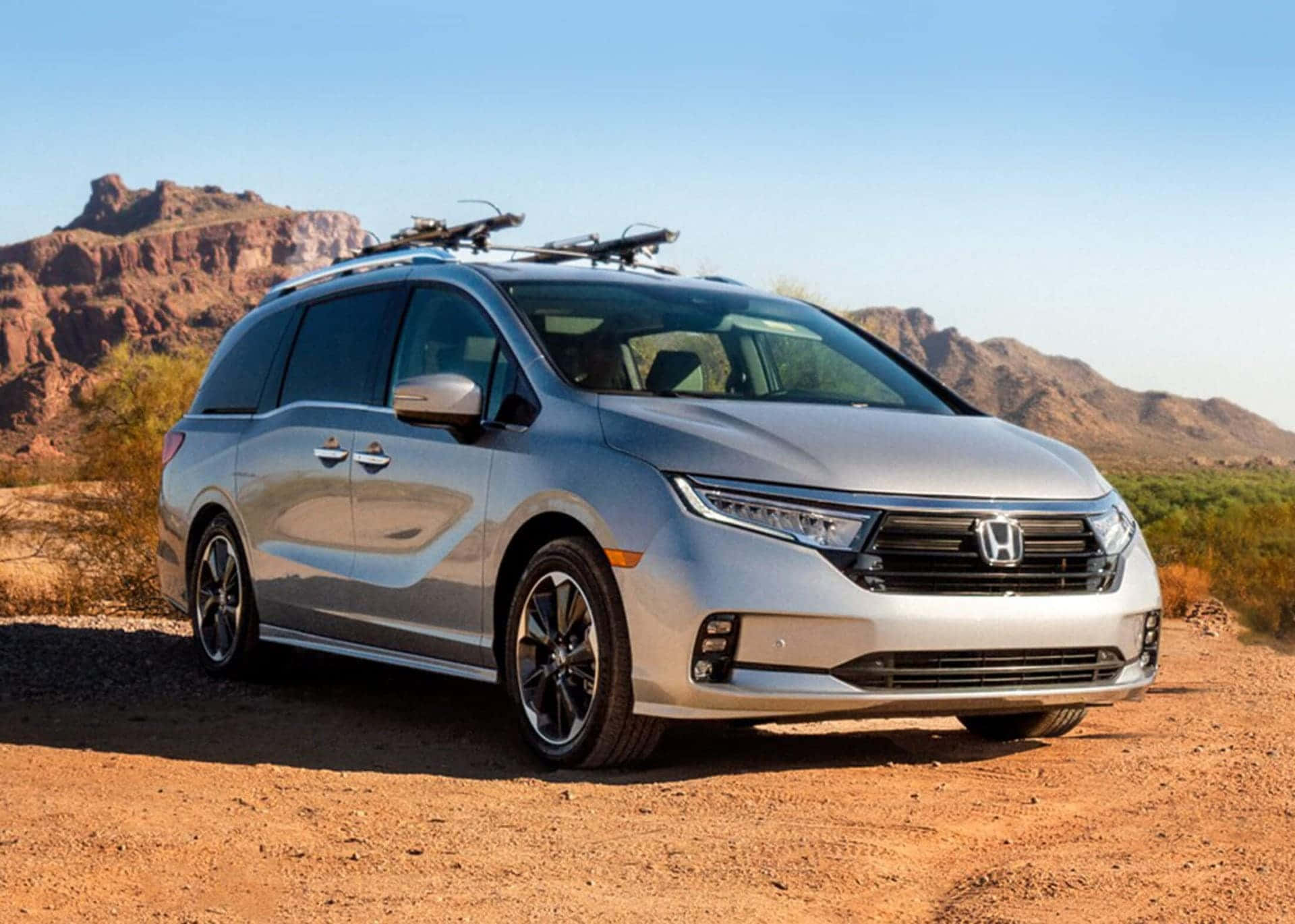Honda Odyssey 桌布 壁紙 — 一輛銀色本田小型貨車配備車頂行李架，停泊在塵土飛揚的沙漠道路上，背景是紅色岩石地貌，天空清澈湛藍。