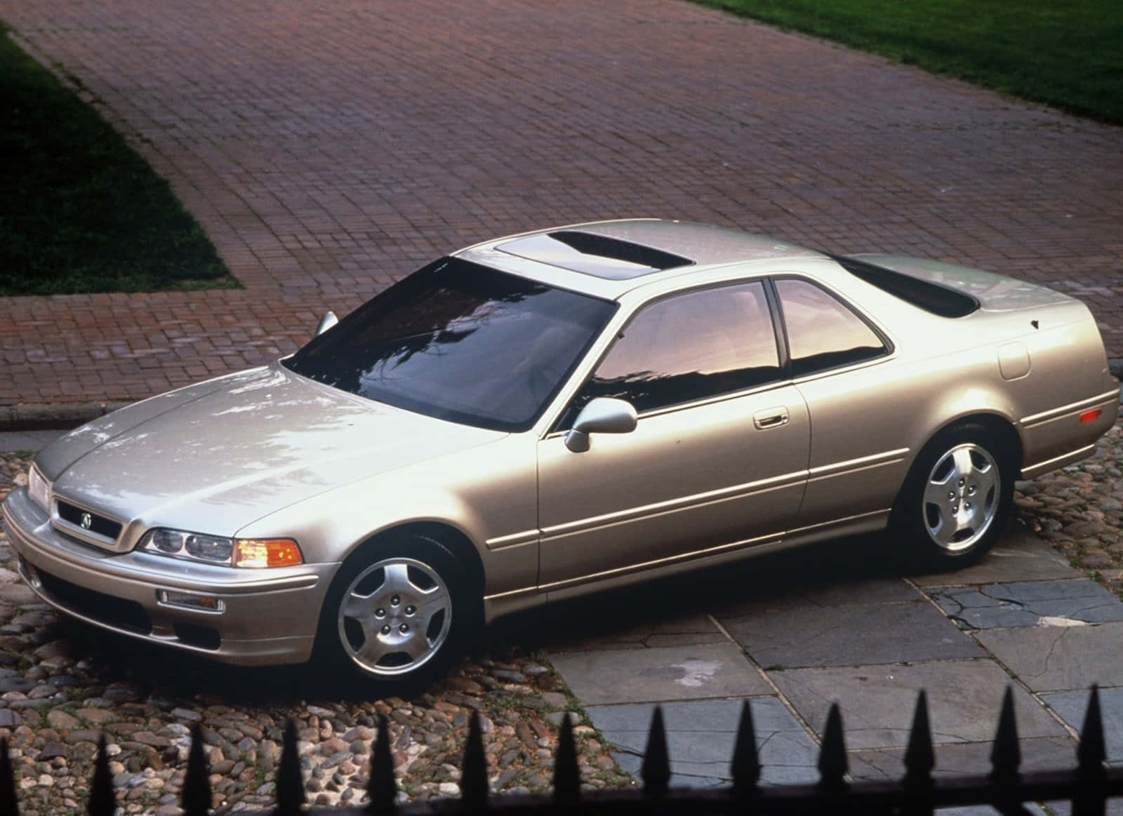 Acura Legend 桌布 壁紙 — 一輛銀色本田轎跑車停放在鵝卵石車道上，旁邊是磚砌小路，前景有黑色鐵欄杆。