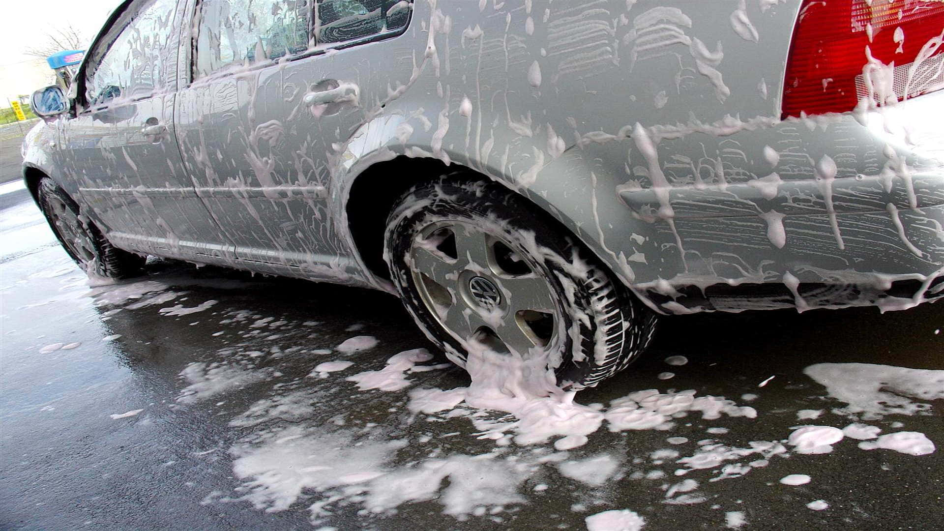 洗車 桌布 壁紙 — 一輛銀色汽車被厚厚的白色肥皂泡覆蓋，正在潮濕的路面上清洗，泡沫從車輪滴落。