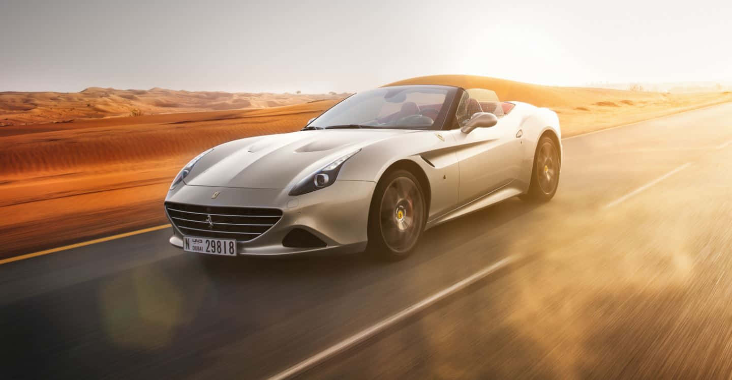 Ferrari California T 桌布 壁紙 — 一輛銀色法拉利敞篷跑車在日落時分沿沙漠公路疾馳，在沙丘和模糊的動態上投下金色光芒。