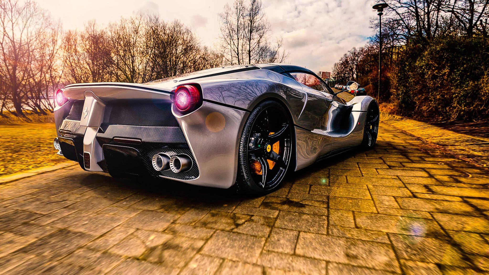 cars 桌布 壁紙 — 一輛銀色法拉利跑車，紅色尾燈發光，停泊在陽光普照的鵝卵石小徑上，周圍是光禿禿的樹木。