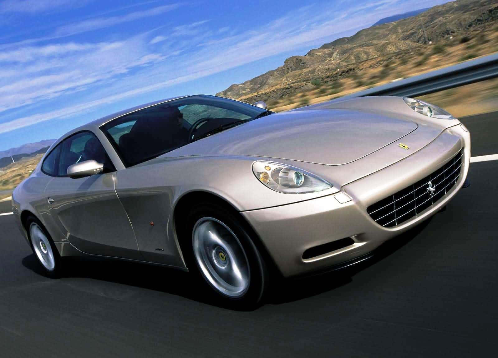 Ferrari 612 Scaglietti 桌布 壁紙 — 一輛銀色法拉利跑車在晴朗藍天白雲下，沿著蜿蜒的山路飛馳。
