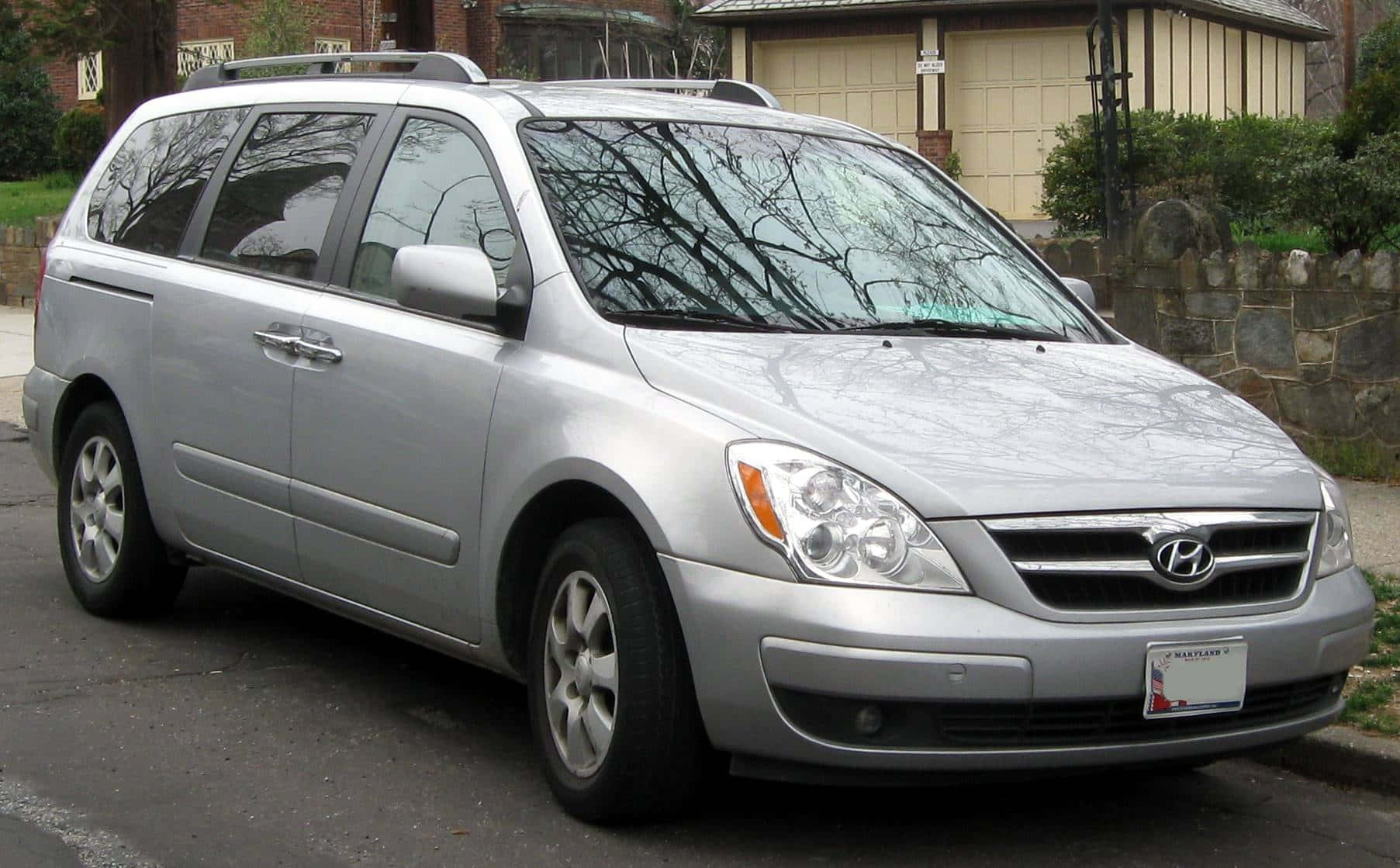 Hyundai Entourage 桌布 壁紙 — 一輛銀色現代Entourage小型貨車停放在住宅街道上，背景有光禿禿的樹木和石牆。